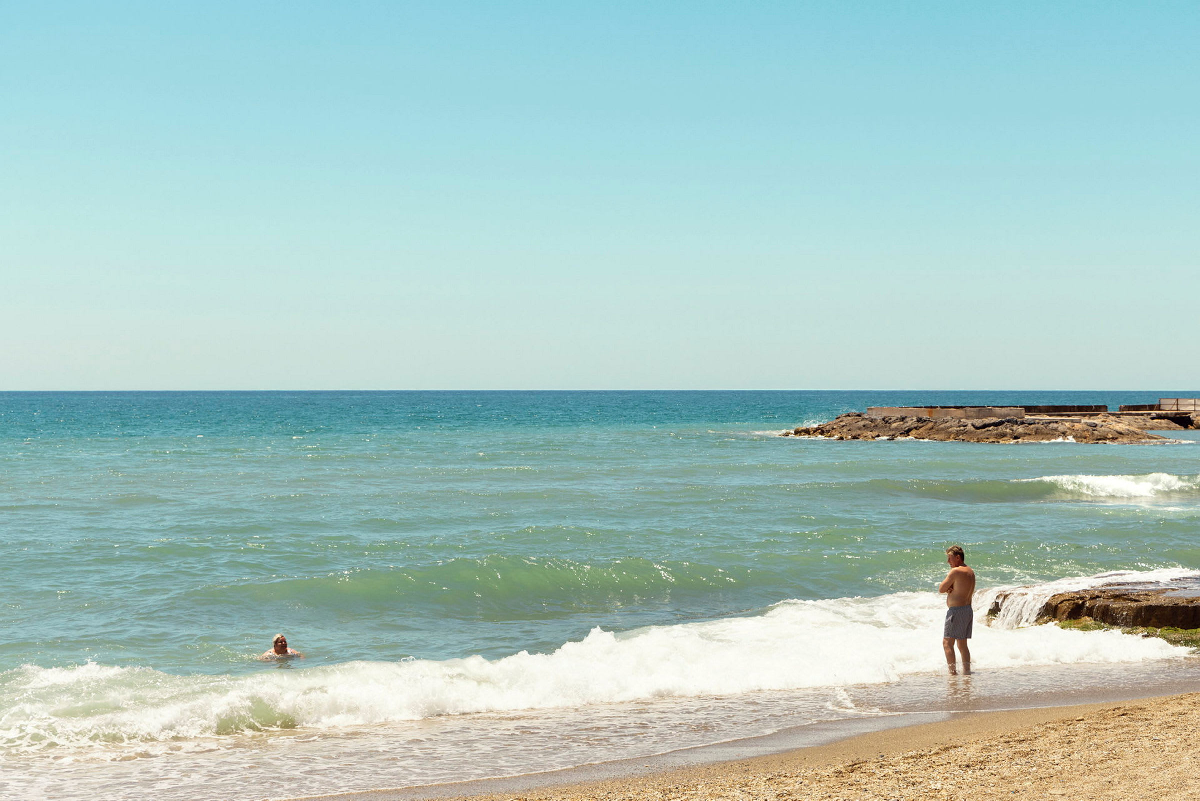 En strand med sand i forgrunnen og bølger som ruller inn. To personer er synlige i bildet, en står ved vannkanten og en svømmer i vannet. (AI-generert tekst)