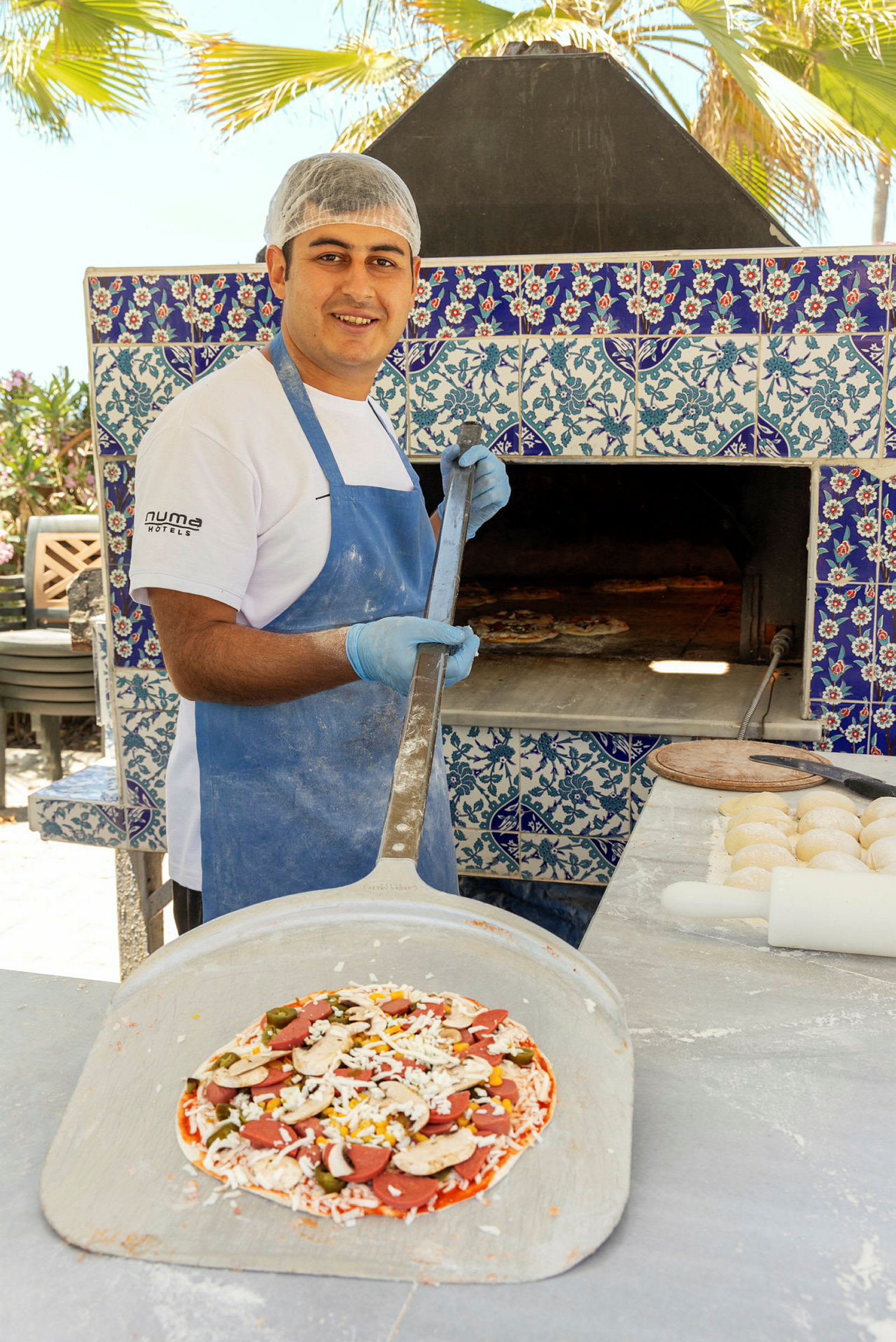 En pizzabaker holder en spade med en ferdiglaget pizza foran en ovn dekorert med blå og hvite fliser. (AI-generert tekst)