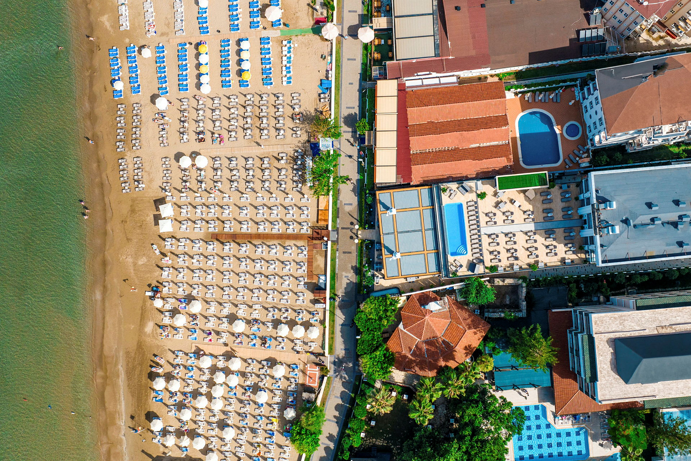 Flyfoto av en strand med solsenger, parasoller og tilstøtende hotellområder med bassenger. (AI-generert tekst)
