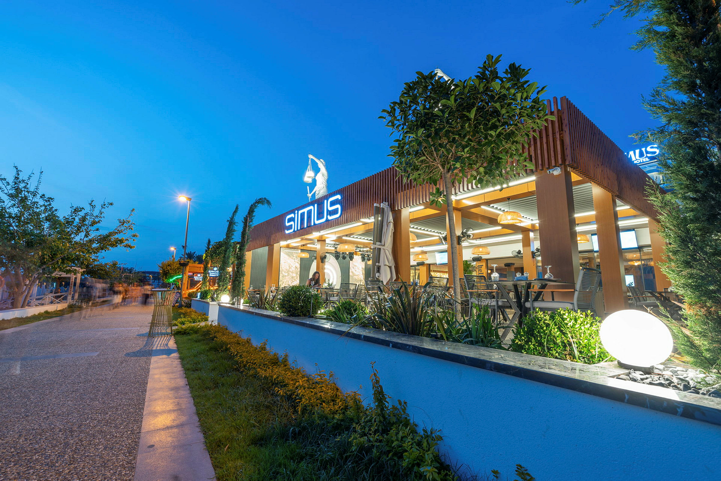 Moderne restaurant med neonskilt, utendørsområde og plantepynt på en kveldssbelyst gate. (AI-generert tekst)