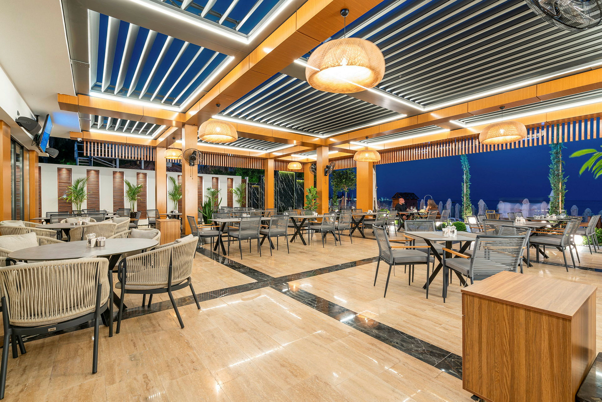 Bilde av en utendørsrestaurant med bord og stoler oppstilt under et moderne tak. (AI-generert tekst)