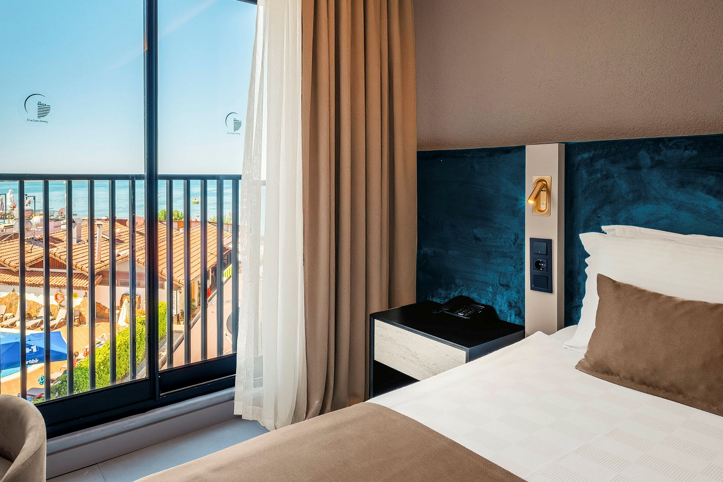 Et hotellrom med havutsikt gjennom store glassvinduer. Innredningen inkluderer en oppredd seng og et nattbord. (AI-generert tekst)