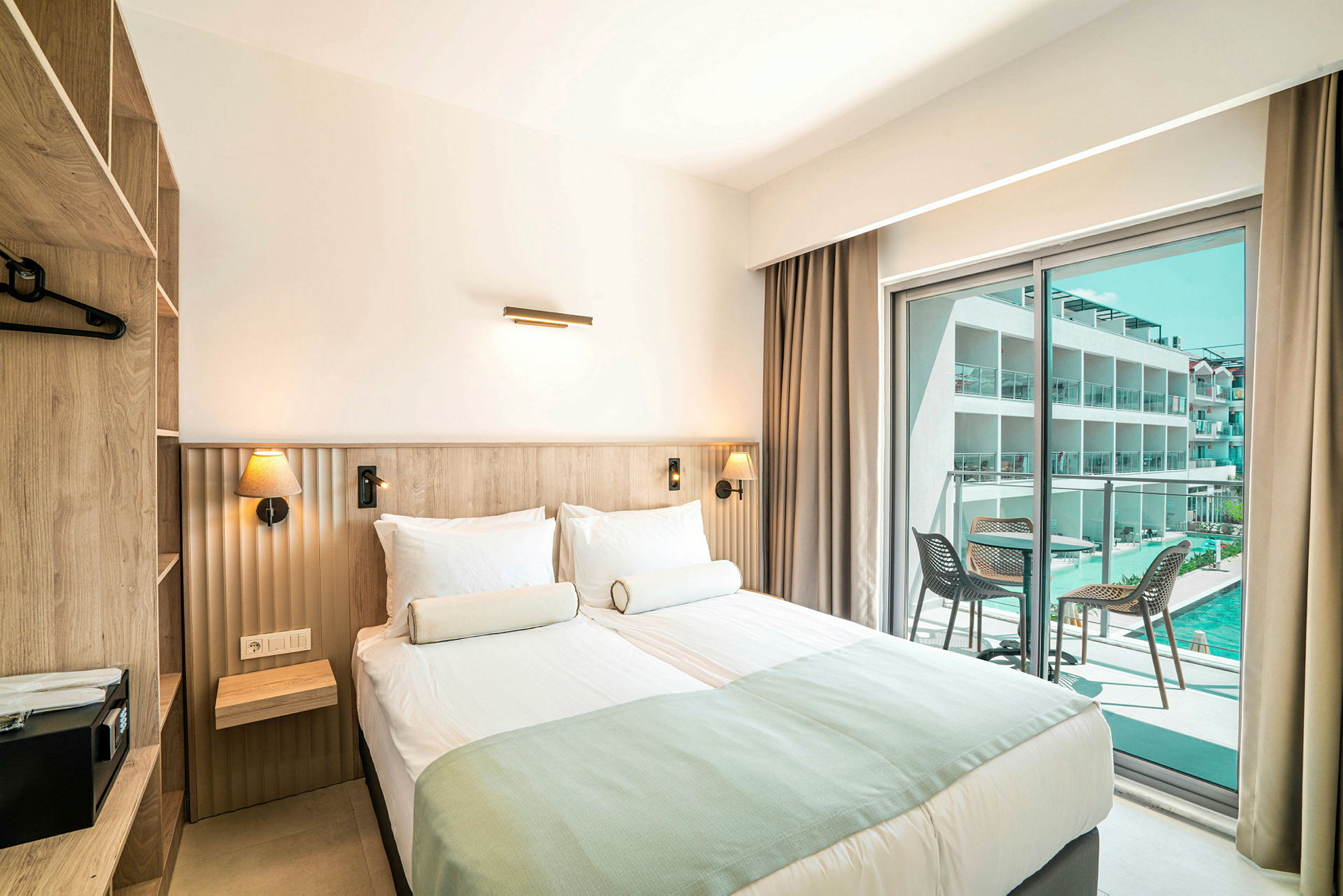 Et hotellrom med dobbeltseng, moderne interiør og glassdører som fører ut til en balkong med utsikt over et basseng og bygninger. (AI-generert tekst)