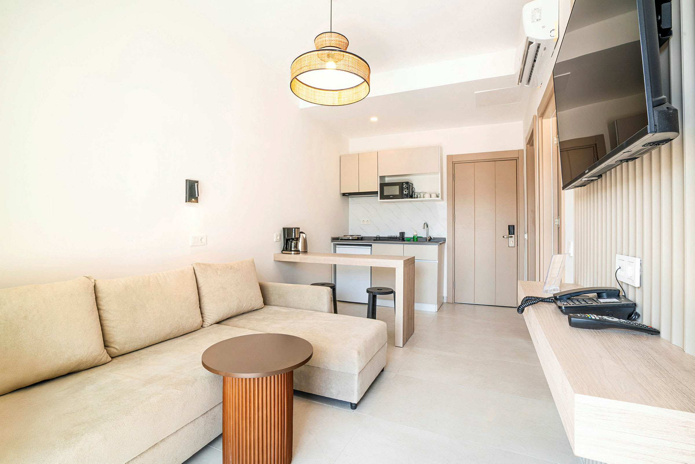 Et hotellrom med en beige sofa, et lite bord, et kjøkken og en veggmontert TV. (AI-generert tekst)