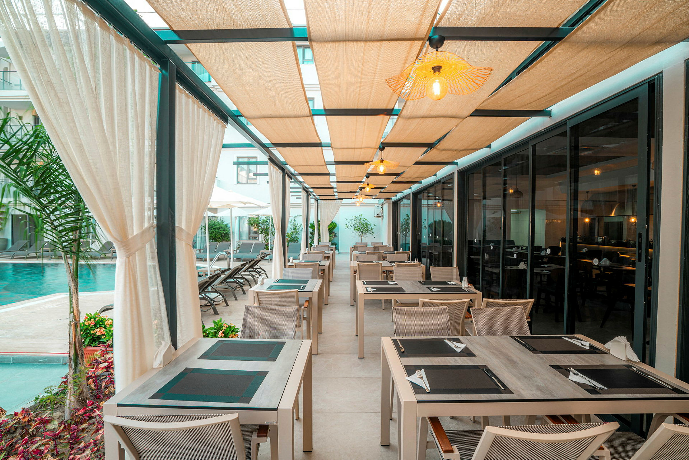 Bilde av en utendørs restaurant med bord og stoler under et tak med lys, ved siden av et basseng. (AI-generert tekst)