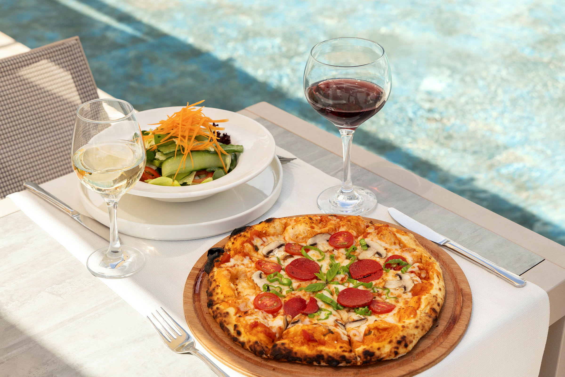 En tallerken med salat og et glass hvitvin ved siden av en pizza med pålegg og et glass rødvin, på et bord med bakgrunn av et basseng. (AI-generert tekst)