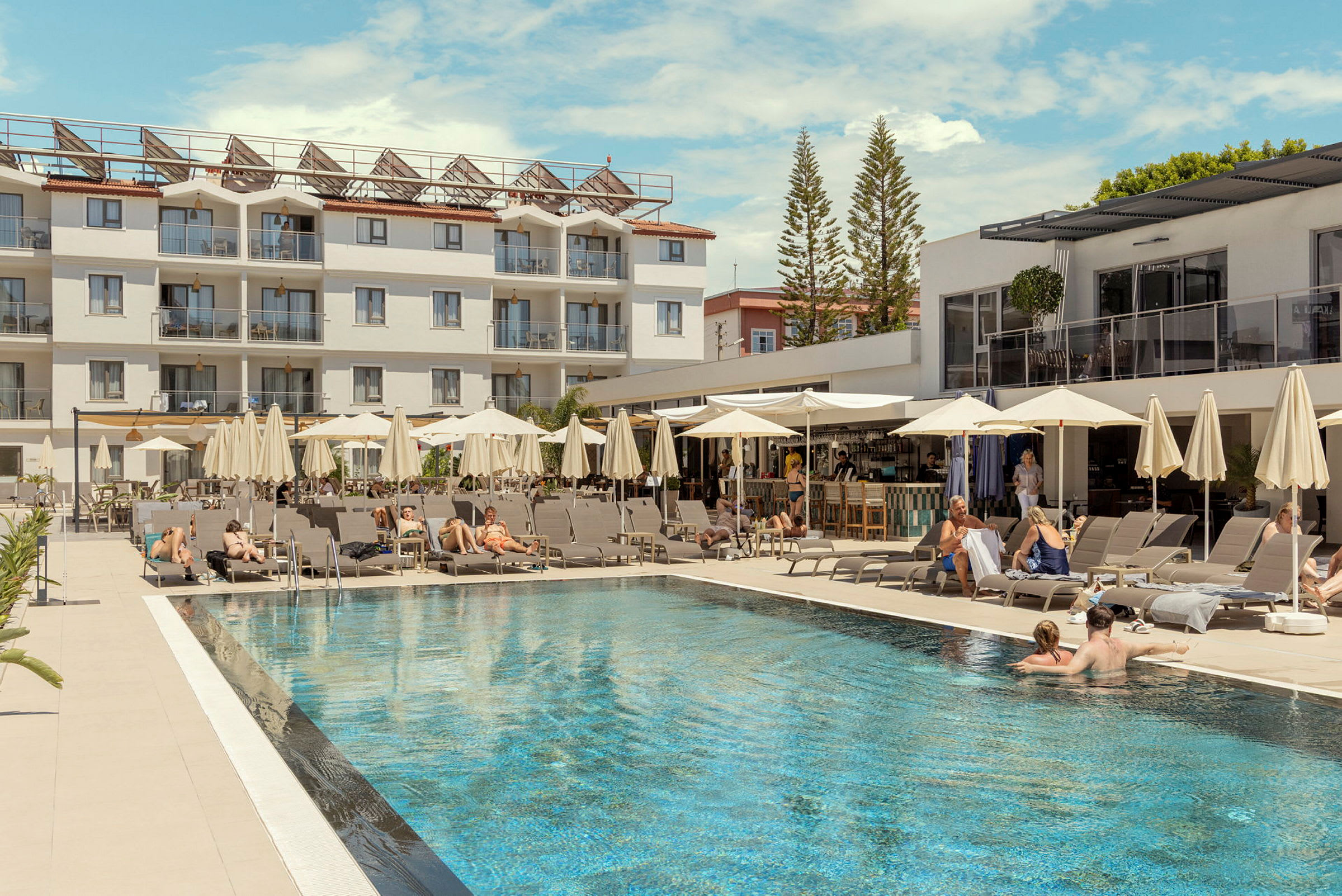 Et hotellbasseng med solsenger og parasoller, omgitt av en hvit bygning og barer. (AI-generert tekst)