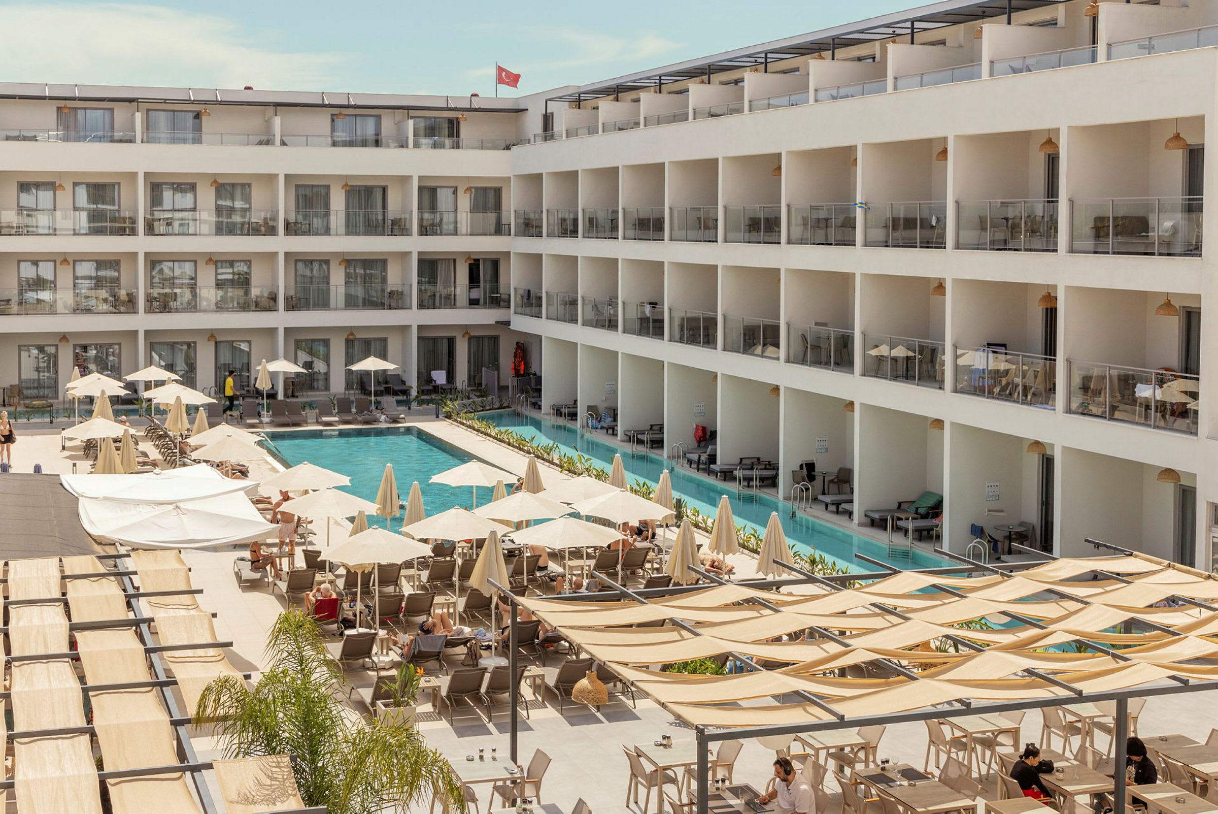 En hotellbygning med et basseng i midten samt solsenger og parasoller rundt. (AI-generert tekst)