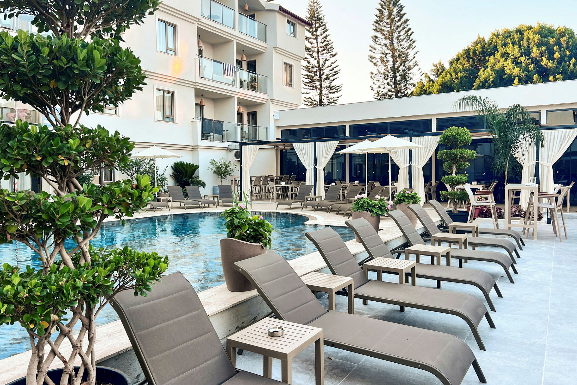 Bassengområde med solsenger, parasoller og bord samt omkringliggende planter ved et hotell. (AI-generert tekst)