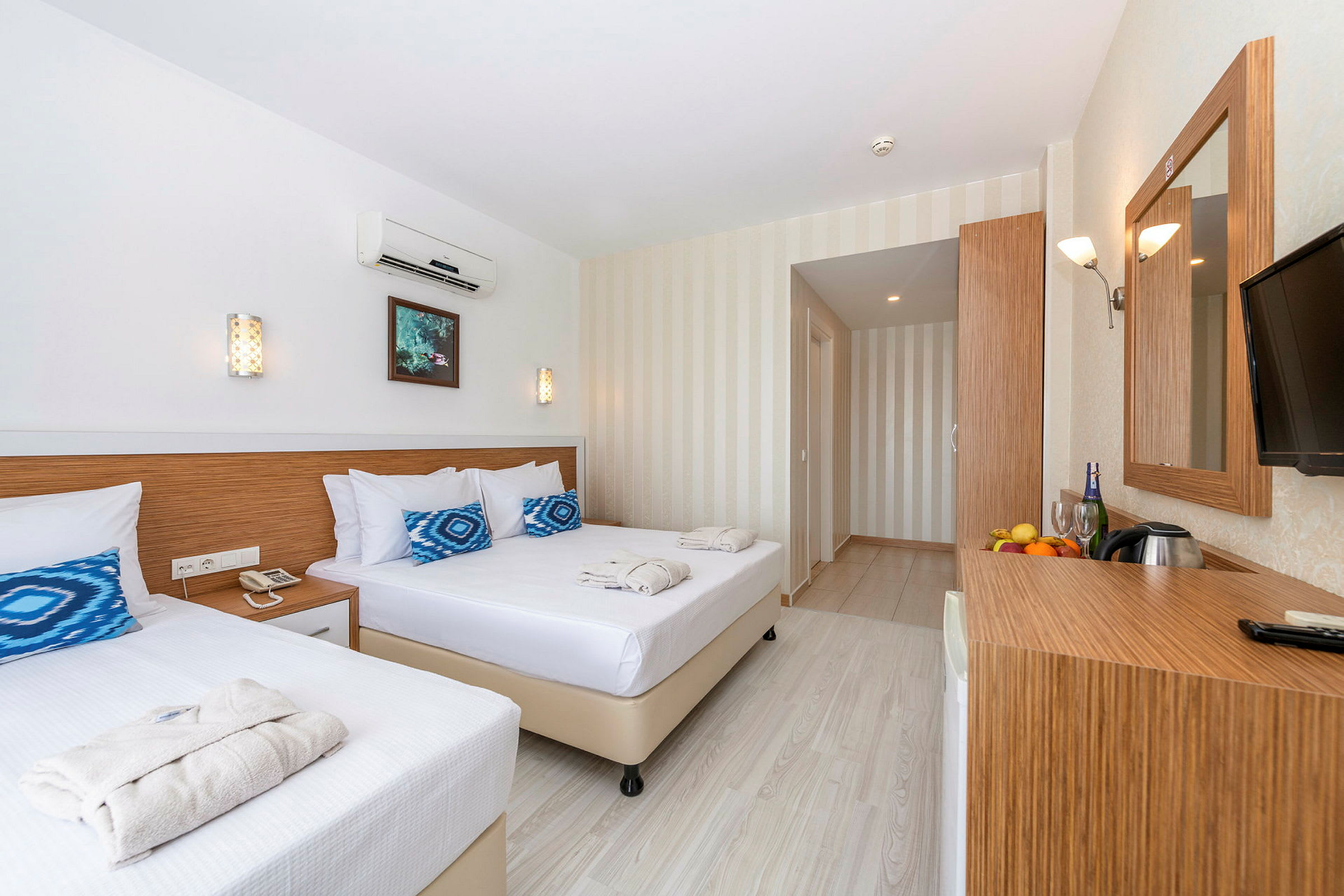 Et lyst og moderne hotellrom med to senger, et speil og et skrivebord med frukt og drikke. (AI-generert tekst)