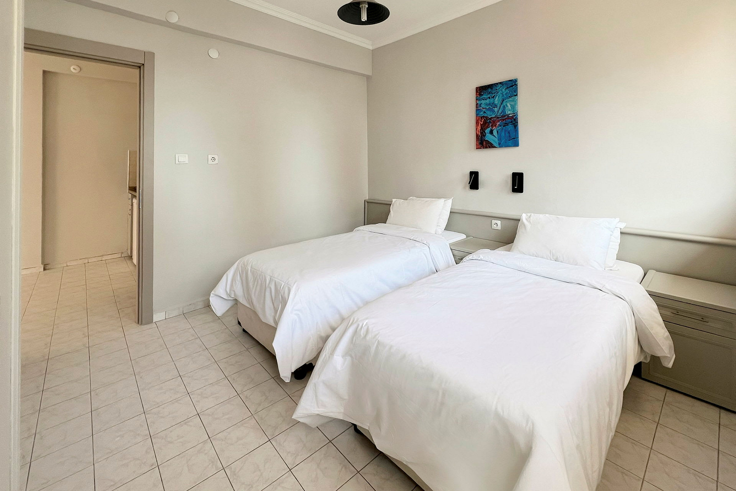 Et hotellrom med to enkeltsenger, hvitt sengetøy og et lyst flislagt gulv. Det er vegglamper og et maleri på veggen over sengene. (AI-generert tekst)
