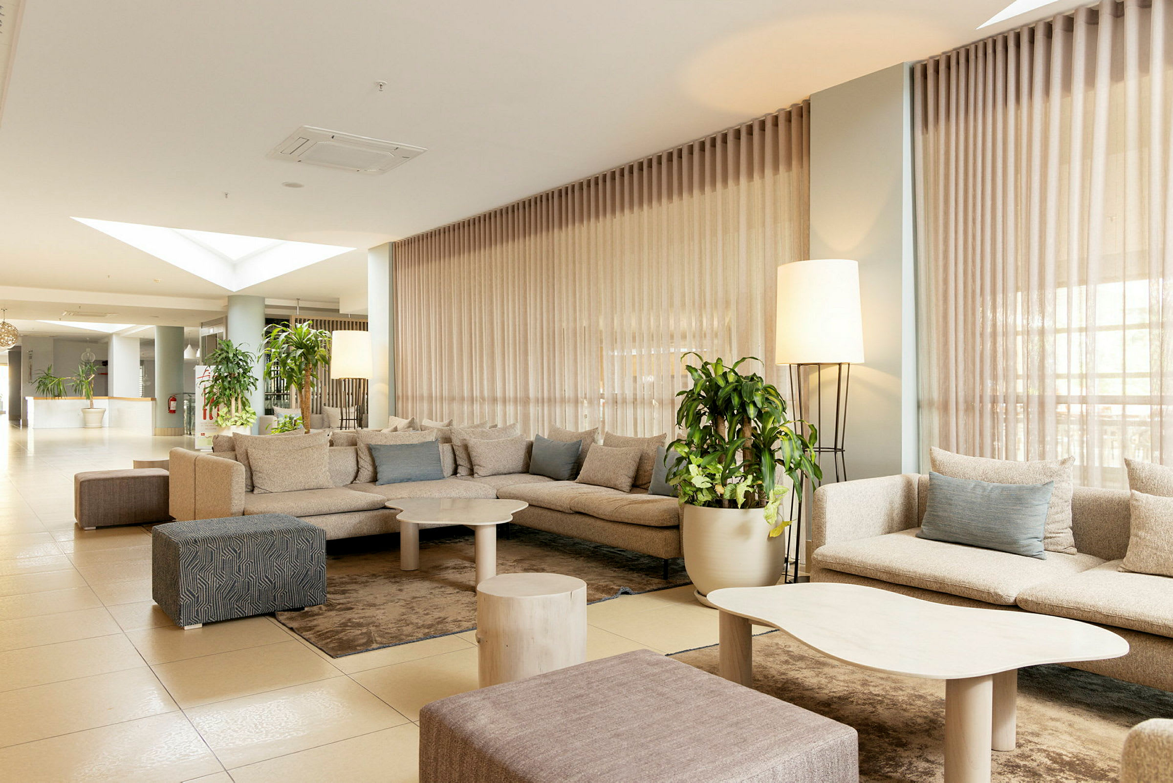 Et hotellrom med moderne design, inneholder en gruppe komfortable beige sofaer, lave bord og grønne planter. (AI-generert tekst)