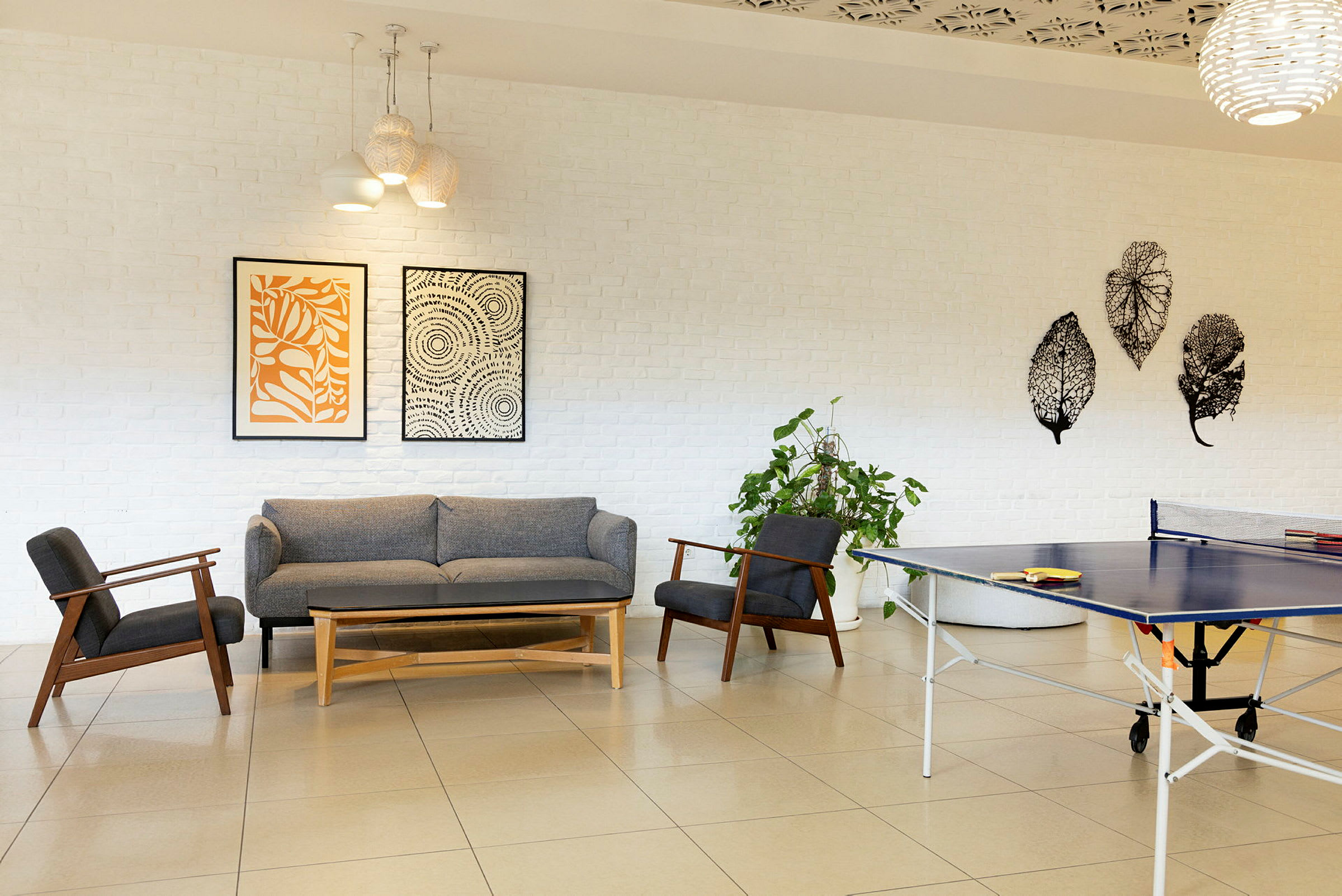En moderne innredet sittegruppe med grå sofa, trestoler og kunst på hvite teglvegger. Et bordtennisbord står nært. (AI-generert tekst)