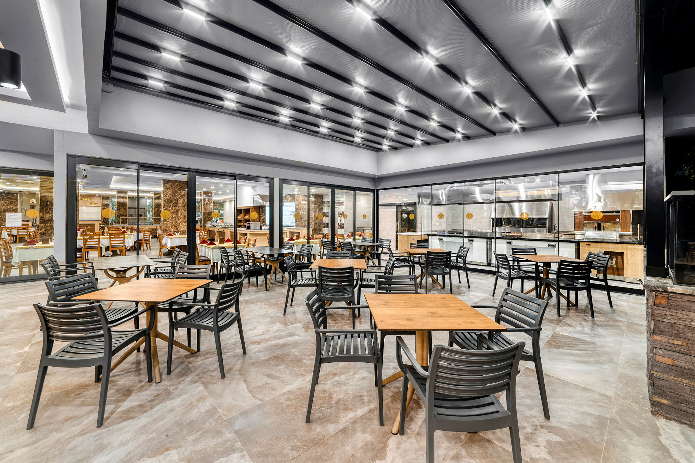 Et moderne restaurantområde med bord og stoler foran en kjøkkenplass med glassvegg. (AI-generert tekst)