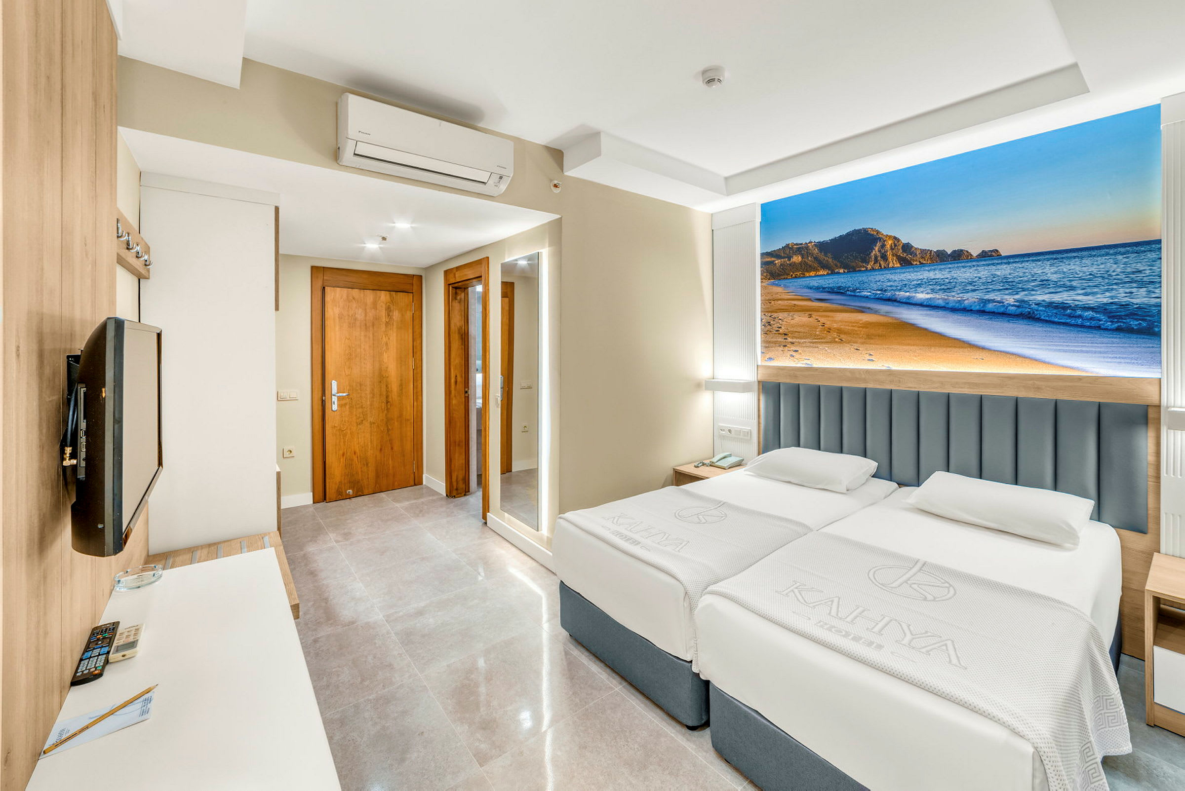 Hotellrom med to senger, en veggmontert TV og et dekorativt bilde av en strand bak sengene. (AI-generert tekst)