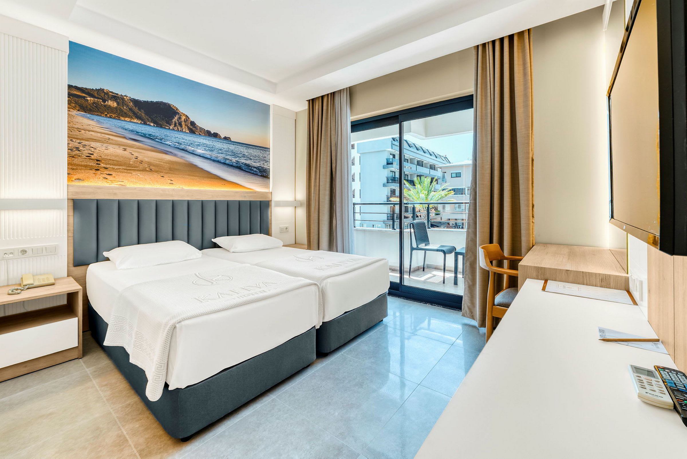 Hotellrom med to enkeltsenger, balkong i bakgrunnen og stort fotografi av en strand på veggen. (AI-generert tekst)