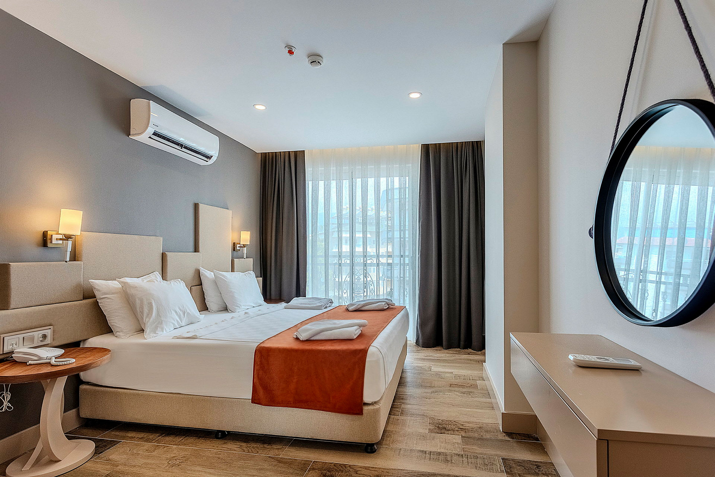 Et hotellrom med en dobbeltseng, nattbord, dekorativt speil på veggen og vindu med gardiner. (AI-generert tekst)