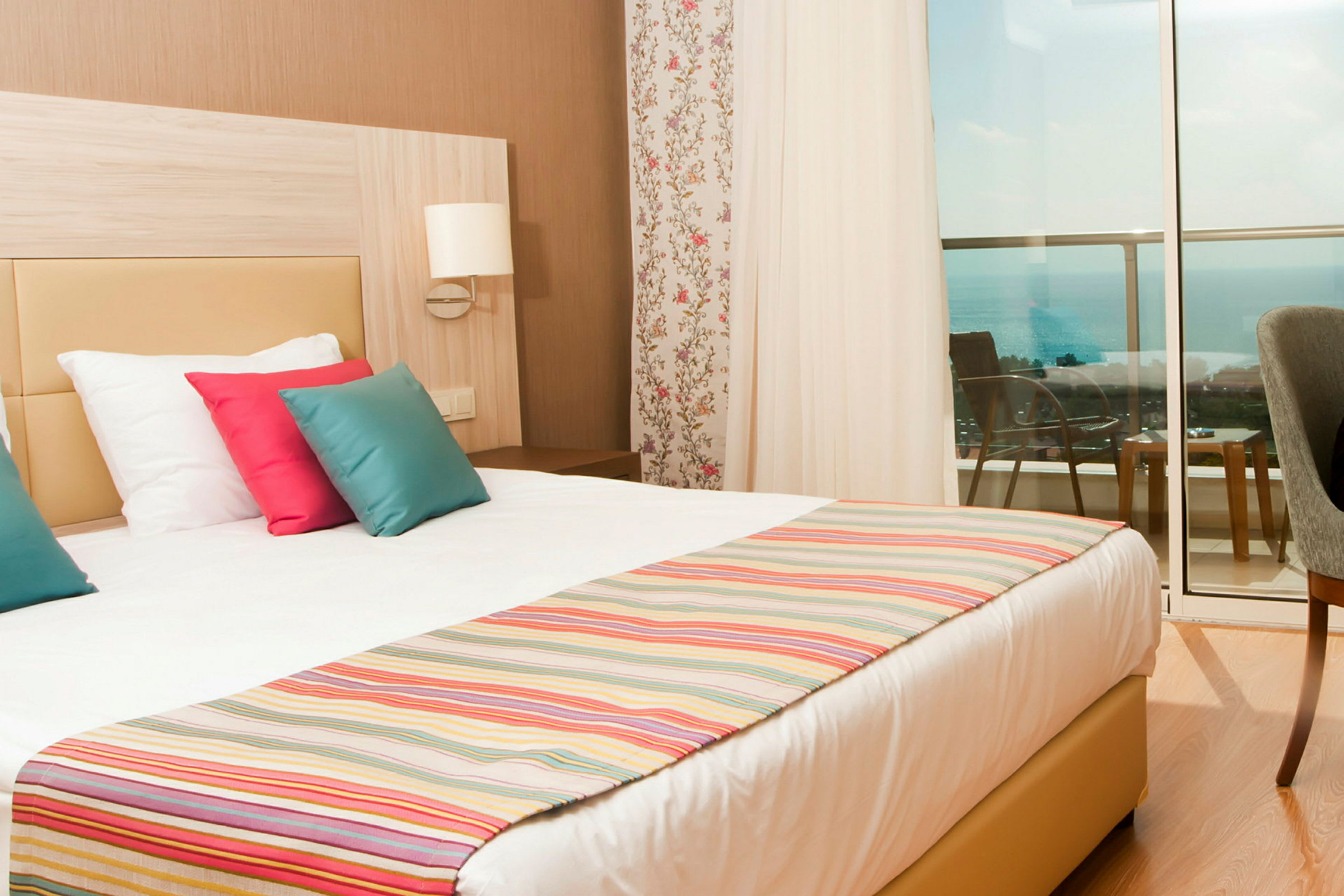 Hotellrom med en dobbeltseng, fargerike puter og utsikt mot havet fra balkongen. (AI-generert tekst)