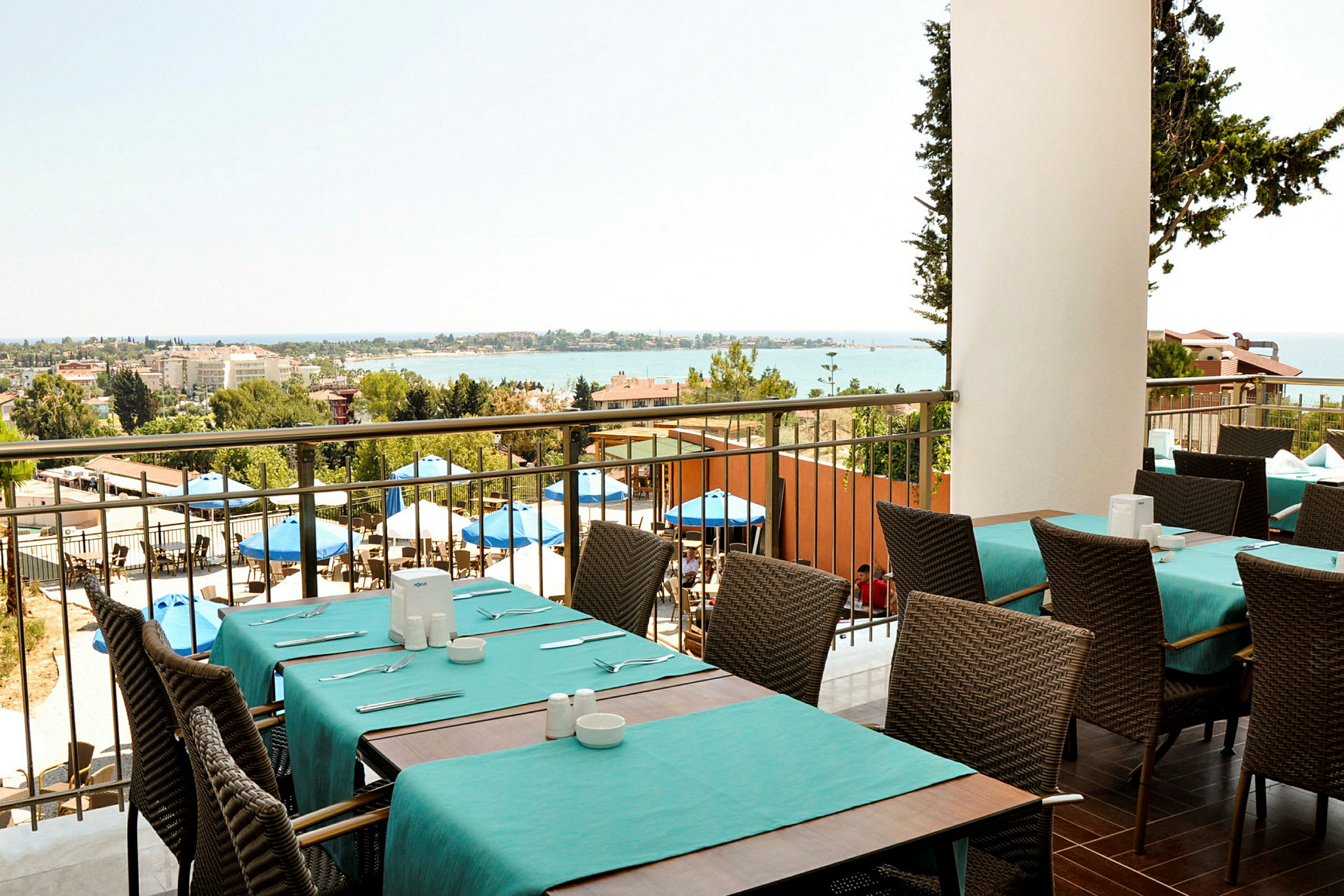Utsikt fra en restaurant med dekkede bord og stoler, uteområde med parasoller og havet i bakgrunnen. (AI-generert tekst)