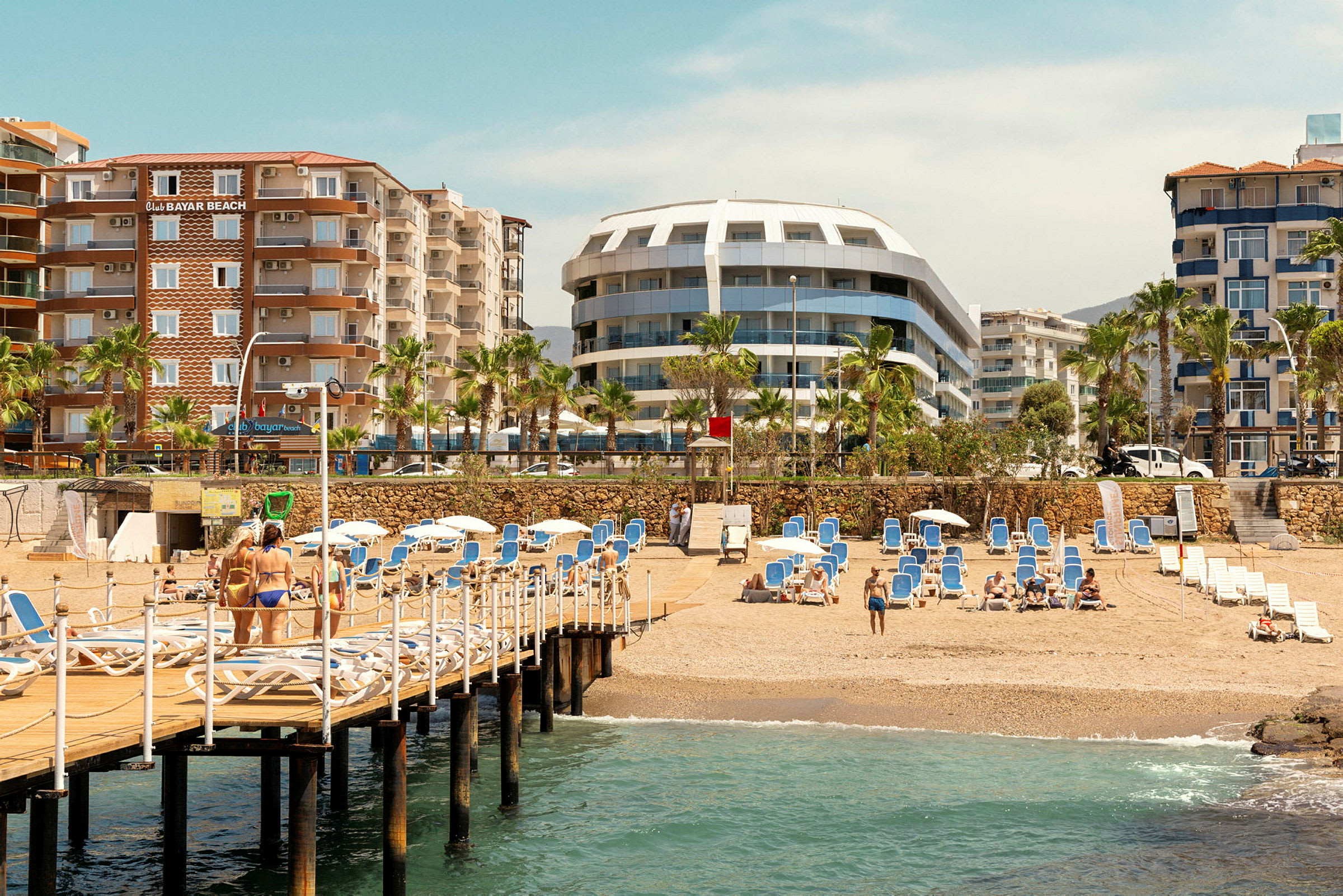 Strand med sand, solsenger, trebrygge og hoteller i bakgrunnen. (AI-generert tekst)