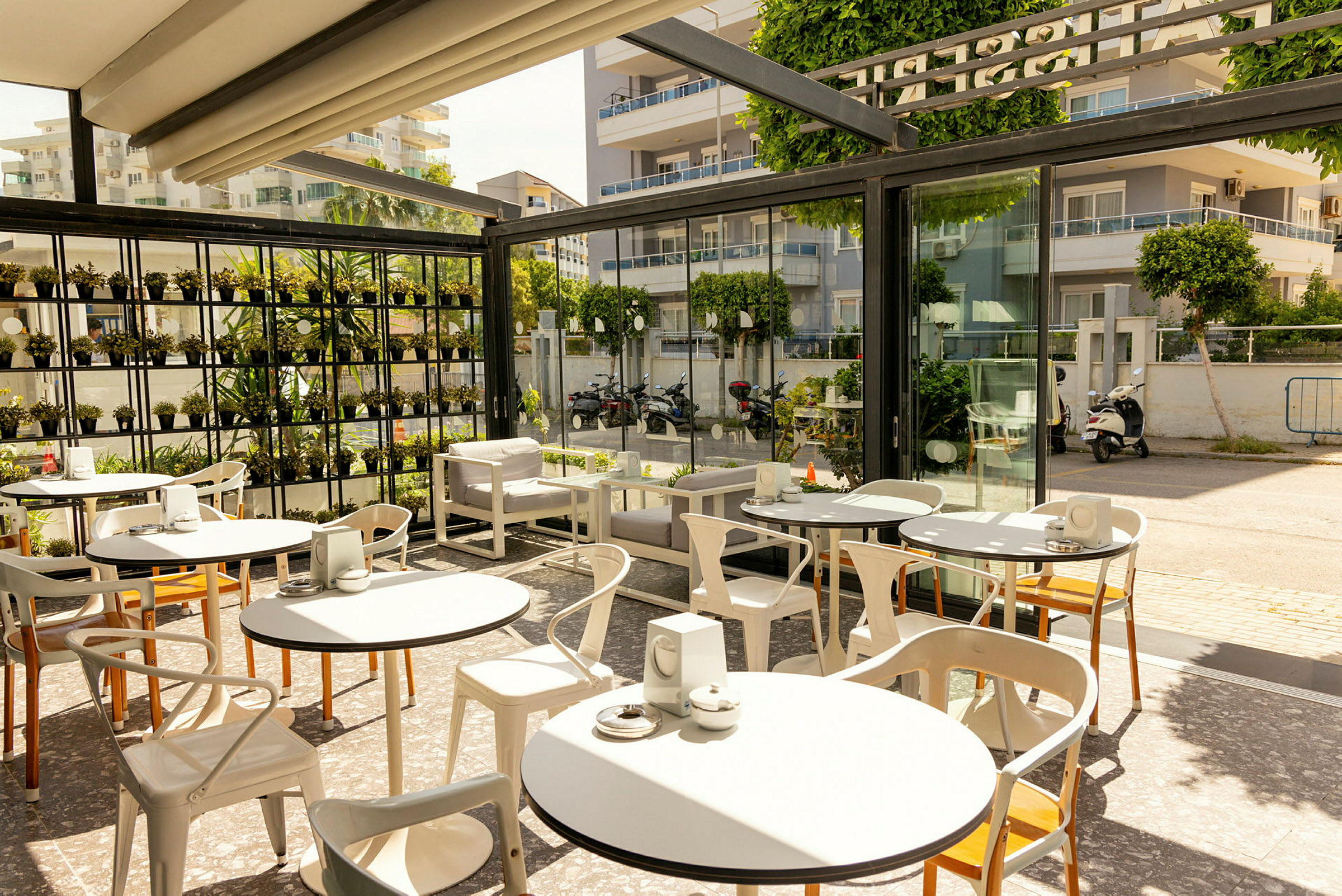 En pen kaféterrasse med bord og stoler, omgitt av glassvegger og grønne planter. (AI-generert tekst)