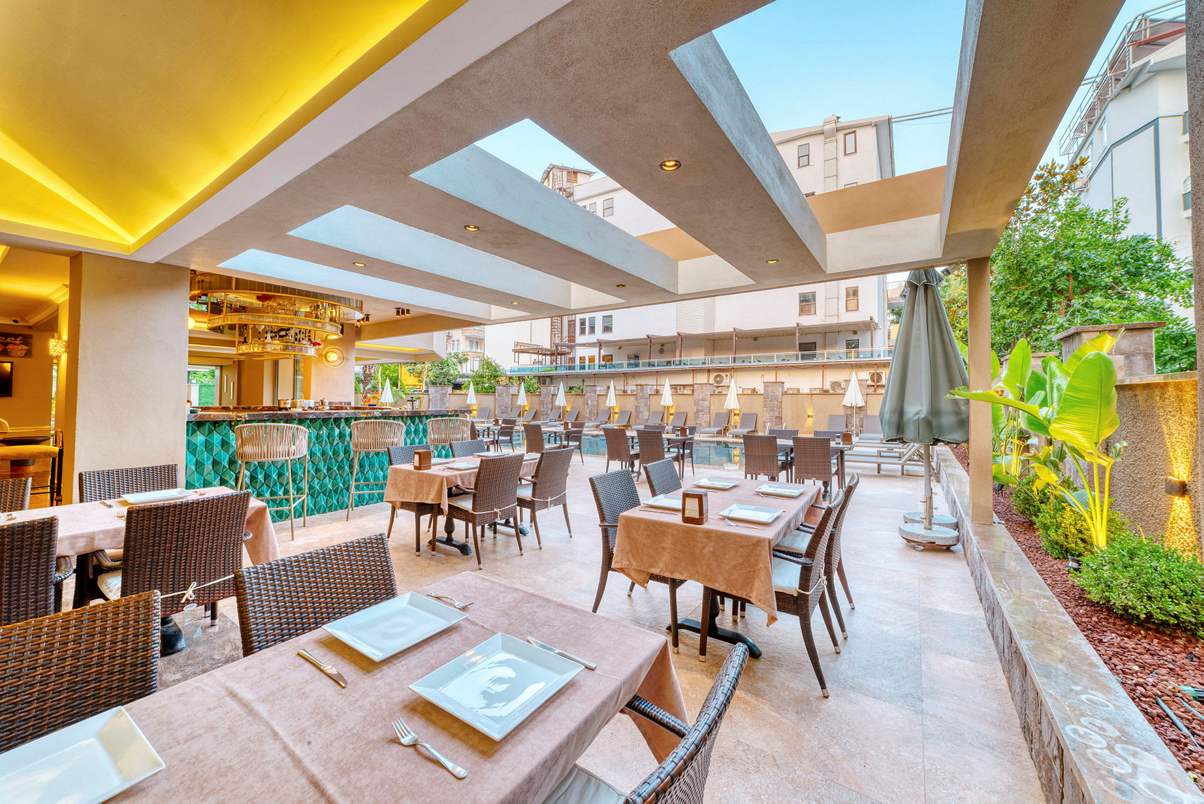 Utendørs restaurant med bord dekket med tallerkener og bestikk, samt grønne planter og basseng i bakgrunnen. (AI-generert tekst)
