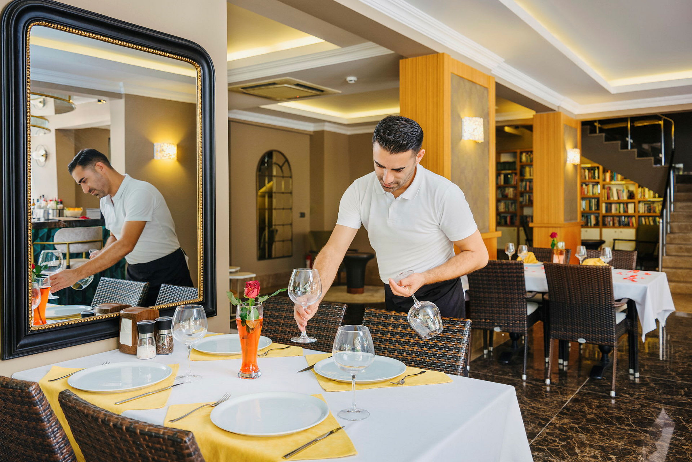 En person setter opp et bord på en restaurant, med hvite tallerkener, glass og gul borddekorasjon. (AI-generert tekst)