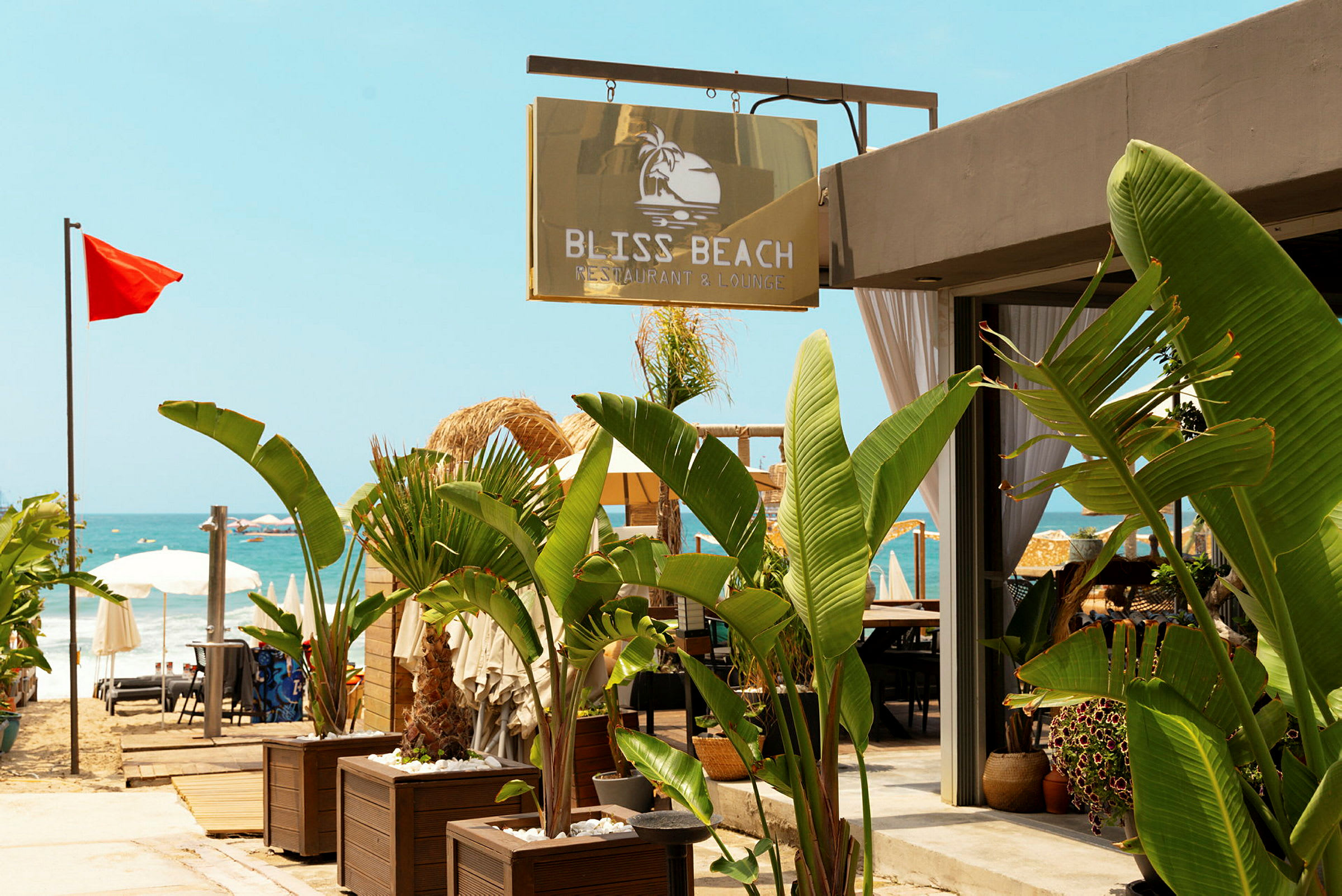 Skilt med teksten Bliss Beach Restaurant & Lounge ved en strand med røde flagg og parasoller. (AI-generert tekst)