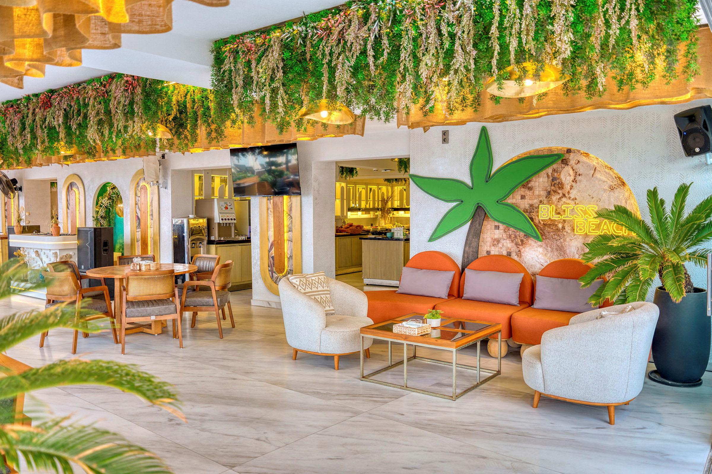 Loungeområde med plantepynt i taket, sitteområde med oransje sofaer, bord og kaffemaskin i bakgrunnen. (AI-generert tekst)