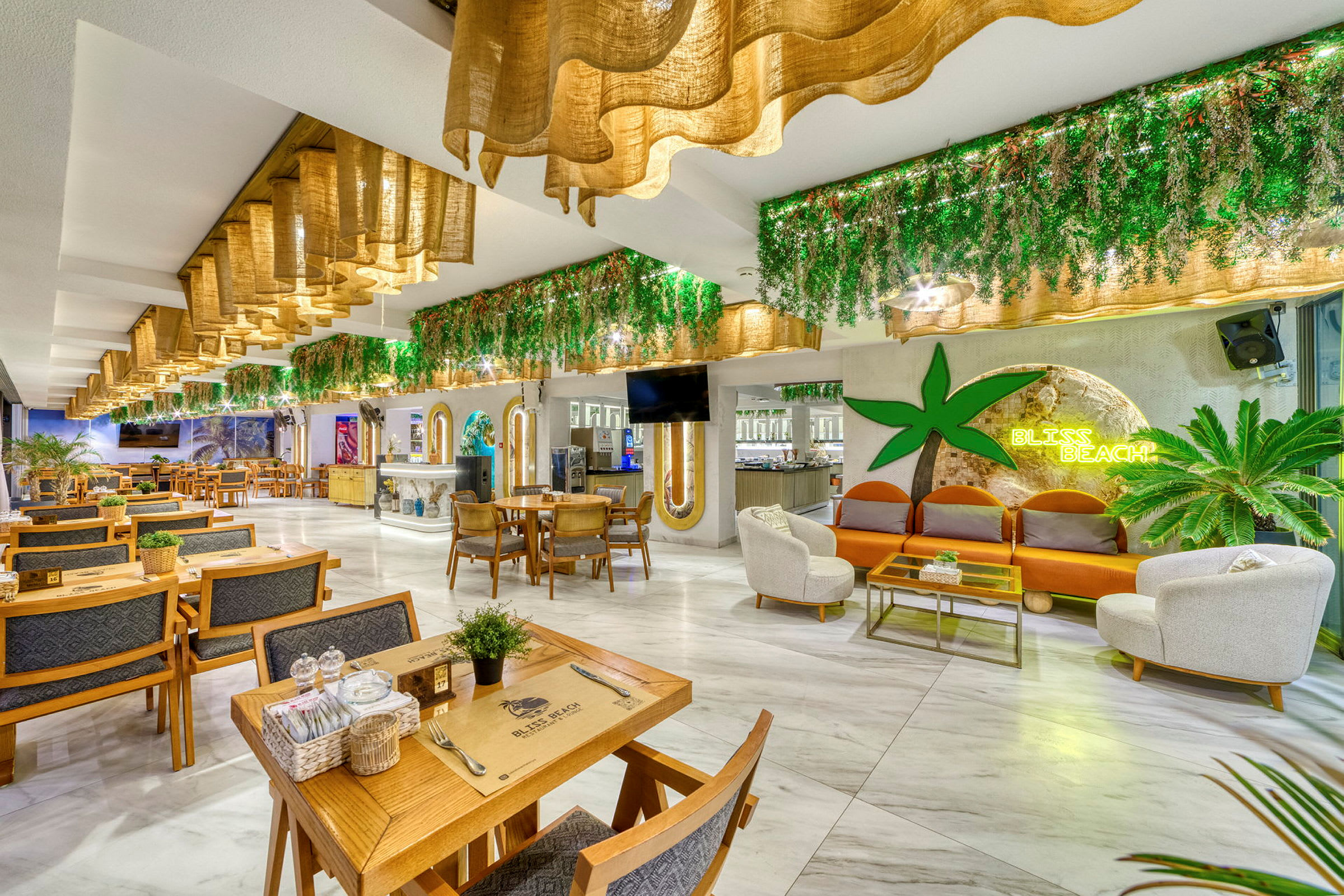 Interiør av en restaurant med sittegrupper, grønne dekorasjoner og et skilt som lyser med teksten Bliss Beach. (AI-generert tekst)