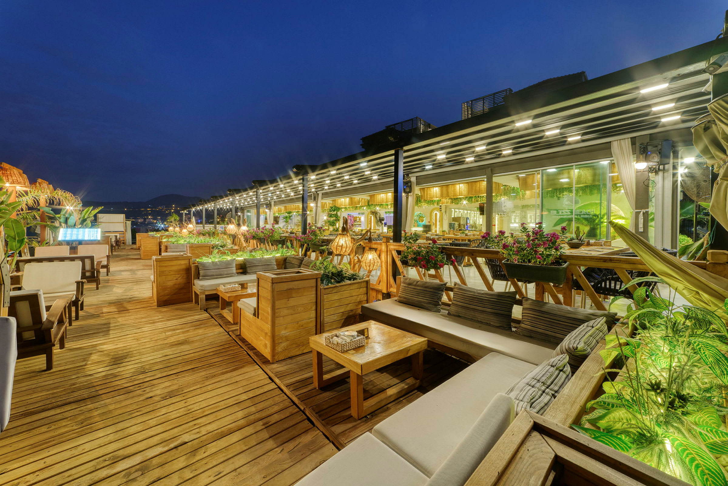 En utendørsrestaurant med treterrasse, sofaer og bord, samt grønne planter og lyskjeder. (AI-generert tekst)