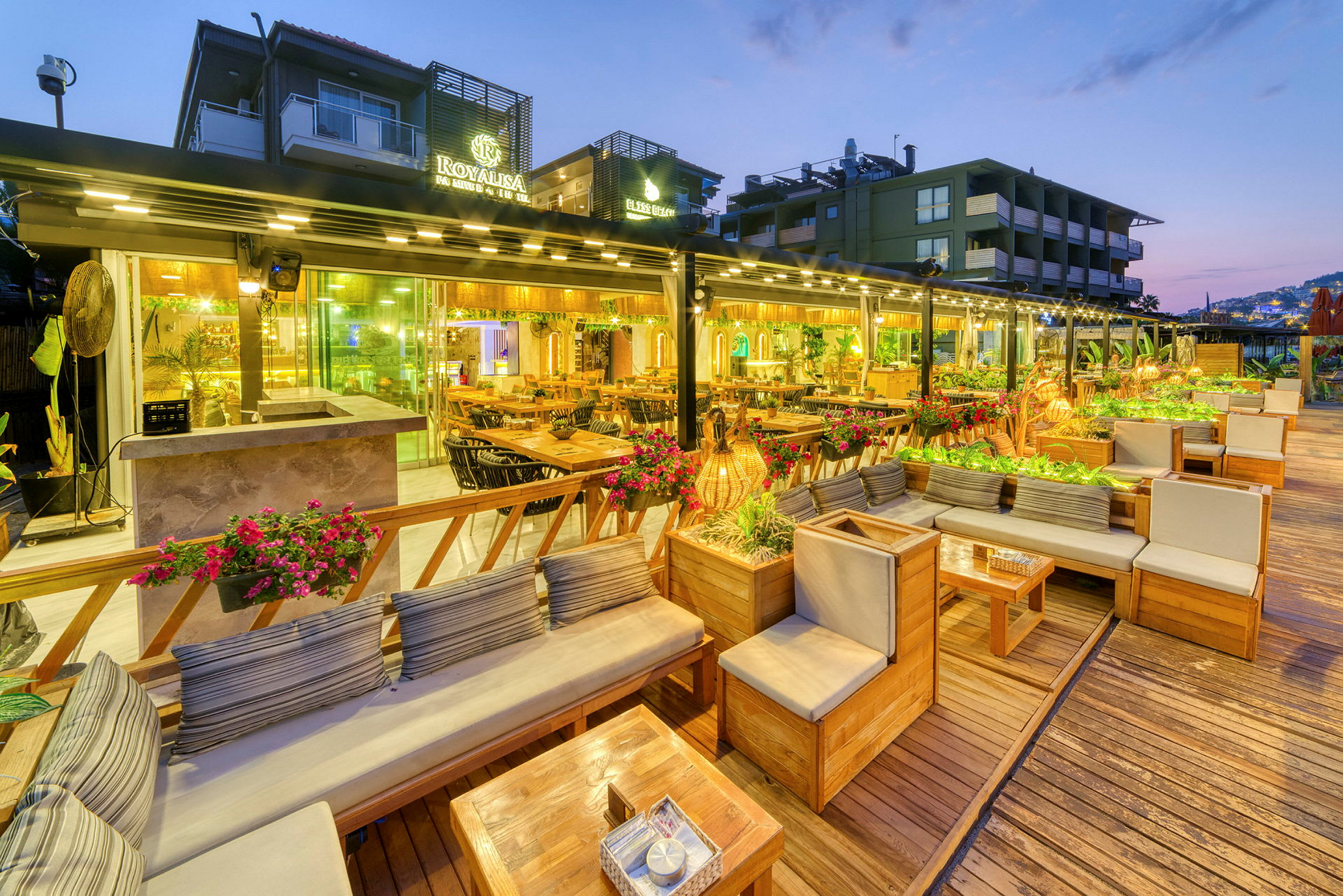 Bilde av en utendørs terrasse med sitteplasser og bord, omgitt av planter og dekorasjoner, opplyst på kvelden. (AI-generert tekst)