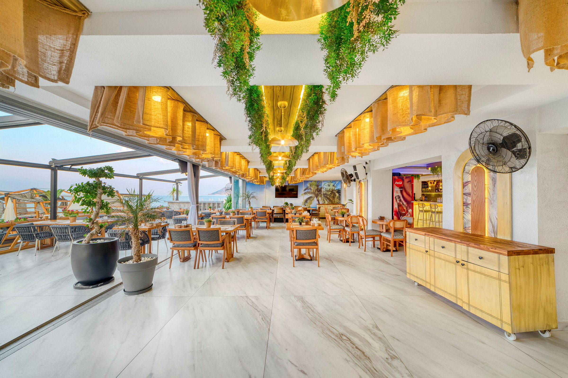 Restaurantinnredning med bord og stoler, grønne dekorasjoner og lyse lamper. (AI-generert tekst)
