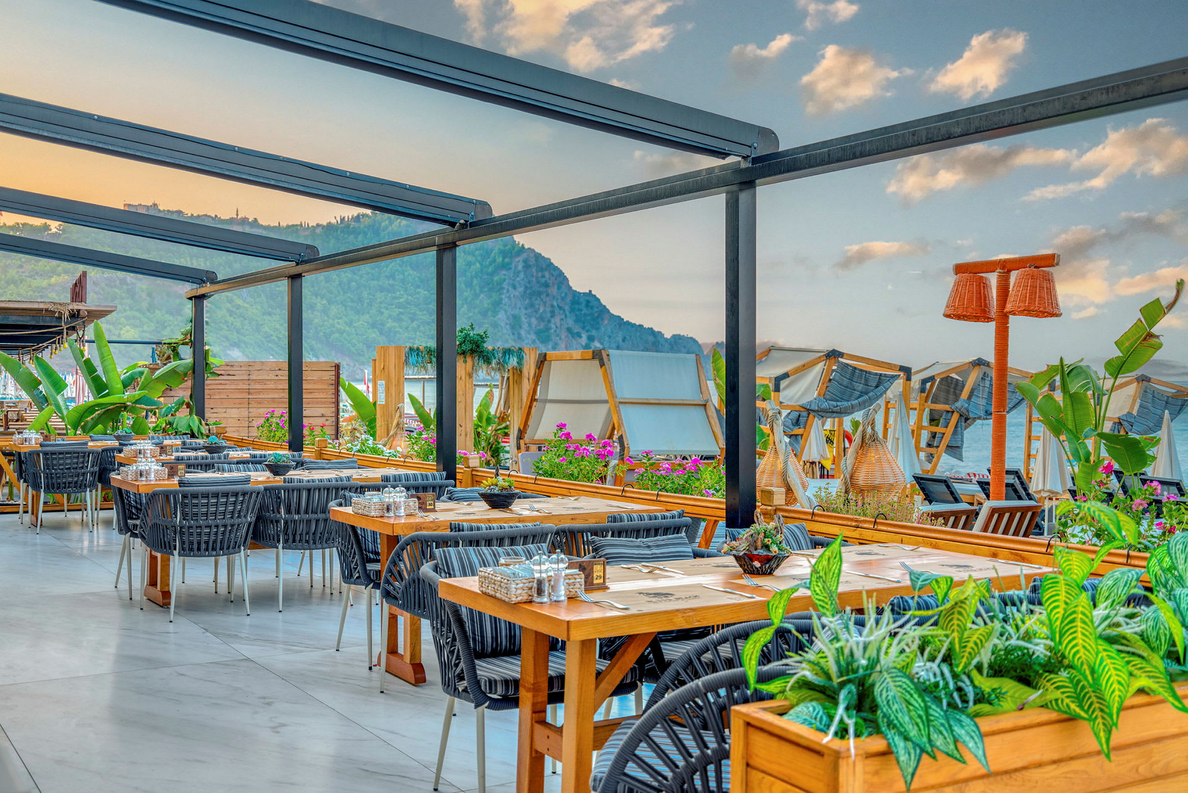 Uterestaurant med bord og stoler, omgitt av planter og med utsikt over havet og fjellene. (AI-generert tekst)