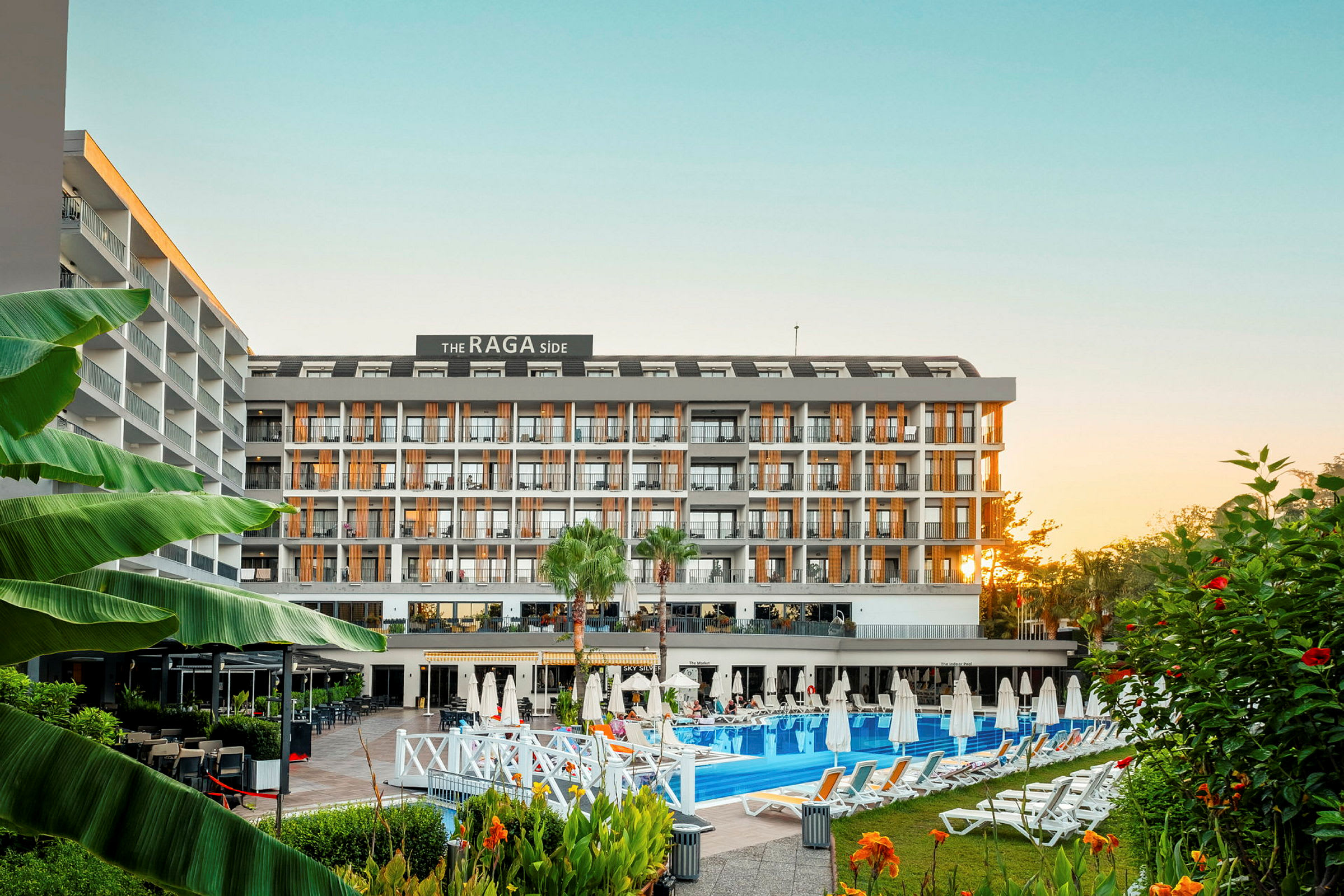 Et fleretasjes hotellkompleks sett fra forsiden, omgitt av basseng, solsenger og vegetasjon. (AI-generert tekst)