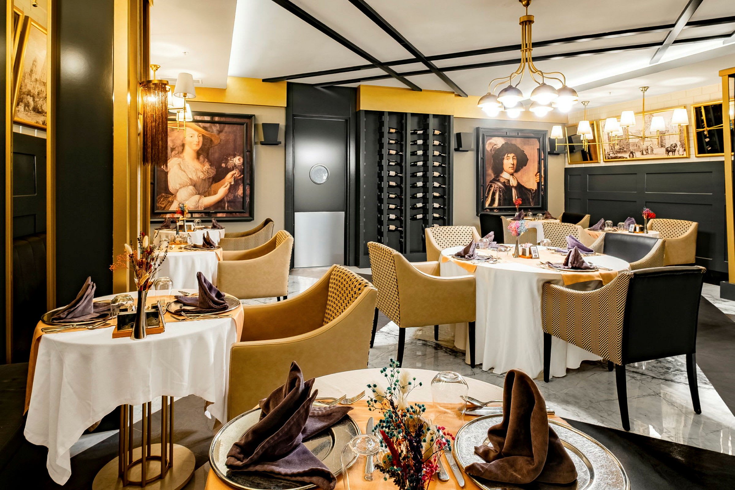 Et elegant innredet restaurantområde med runde bord dekket med hvite duker, omgitt av moderne stoler og dekorasjoner som inkluderer kunstverk på veggene og en vinhylle. (AI-generert tekst)
