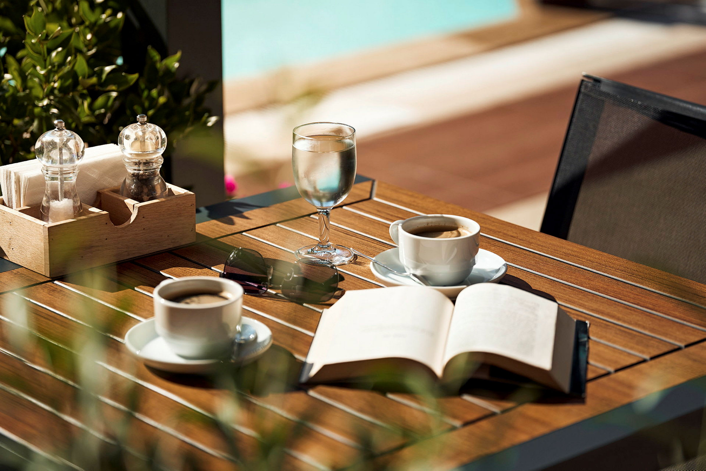 Et bord med kaffekopper, et glass vann, en åpen bok og solbriller. (AI-generert tekst)