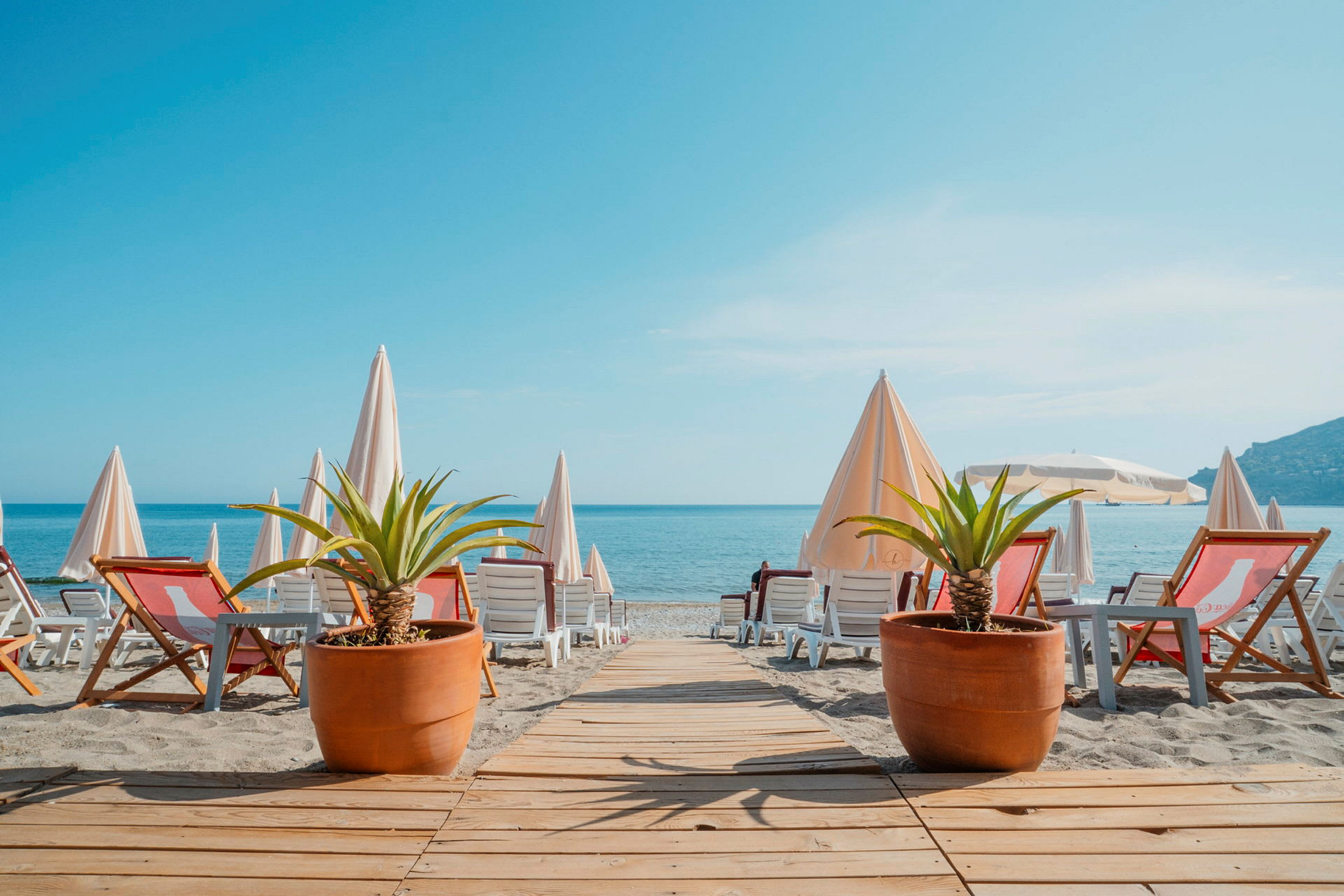 Strand med solsenger, parasoller og to store potteplanter. (AI-generert tekst)