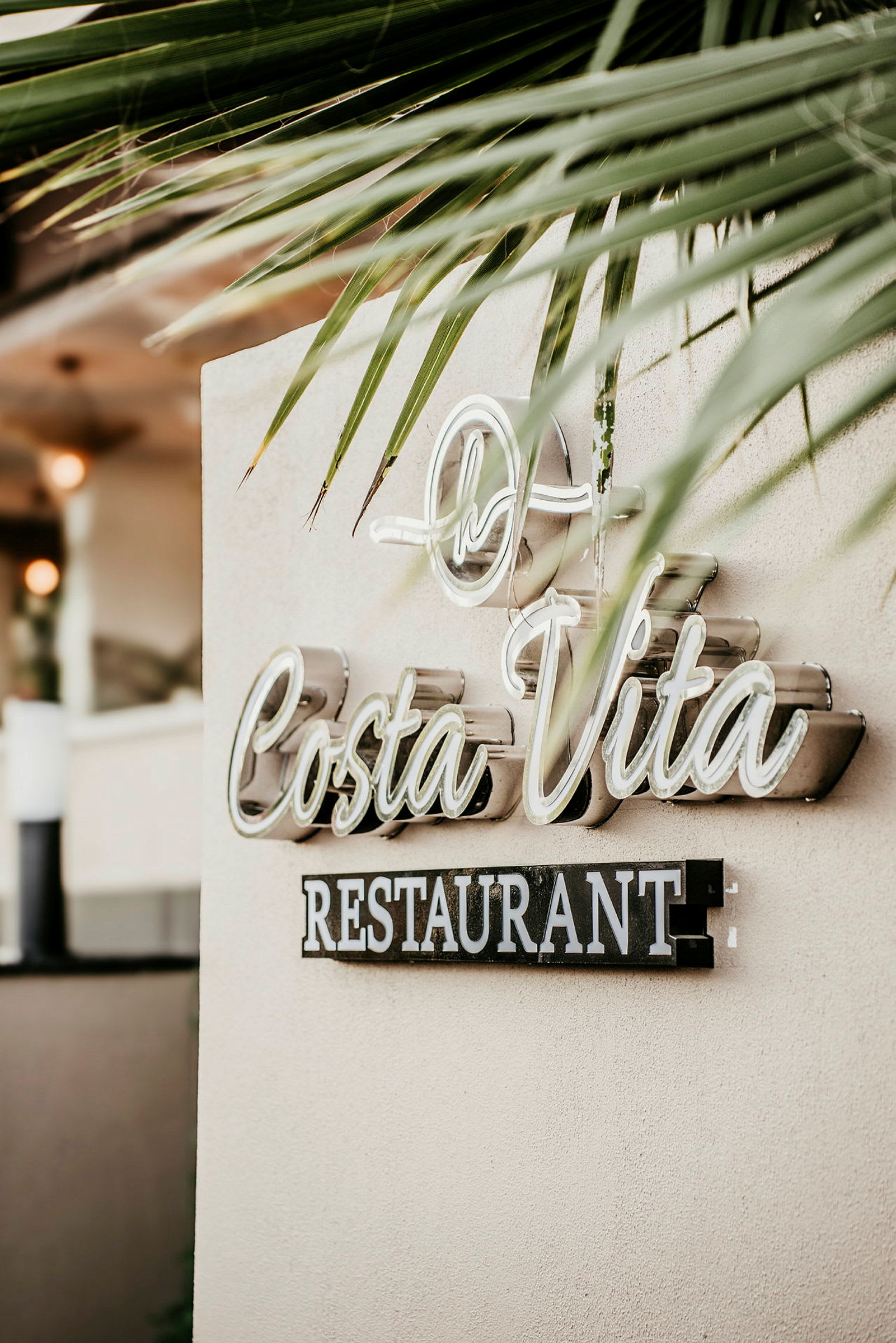 Et skilt med teksten 'Costa Vita Restaurant' på en vegg, omgitt av palmebladdekorasjon. (AI-generert tekst)