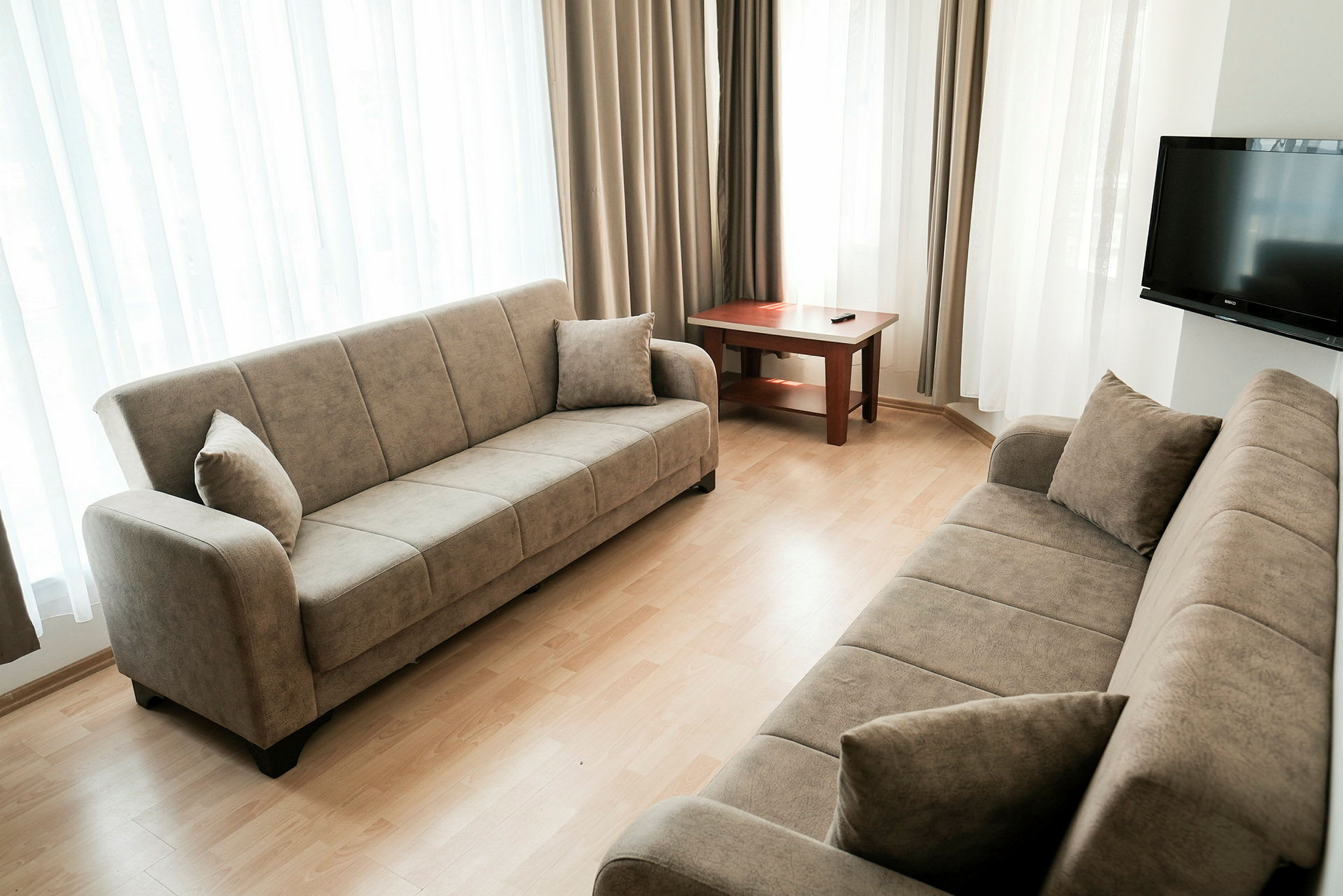 Et stue med to beige sofaer, et trebord og en TV på veggen. Rommet har lyse gardiner ved vinduene. (AI-generert tekst)