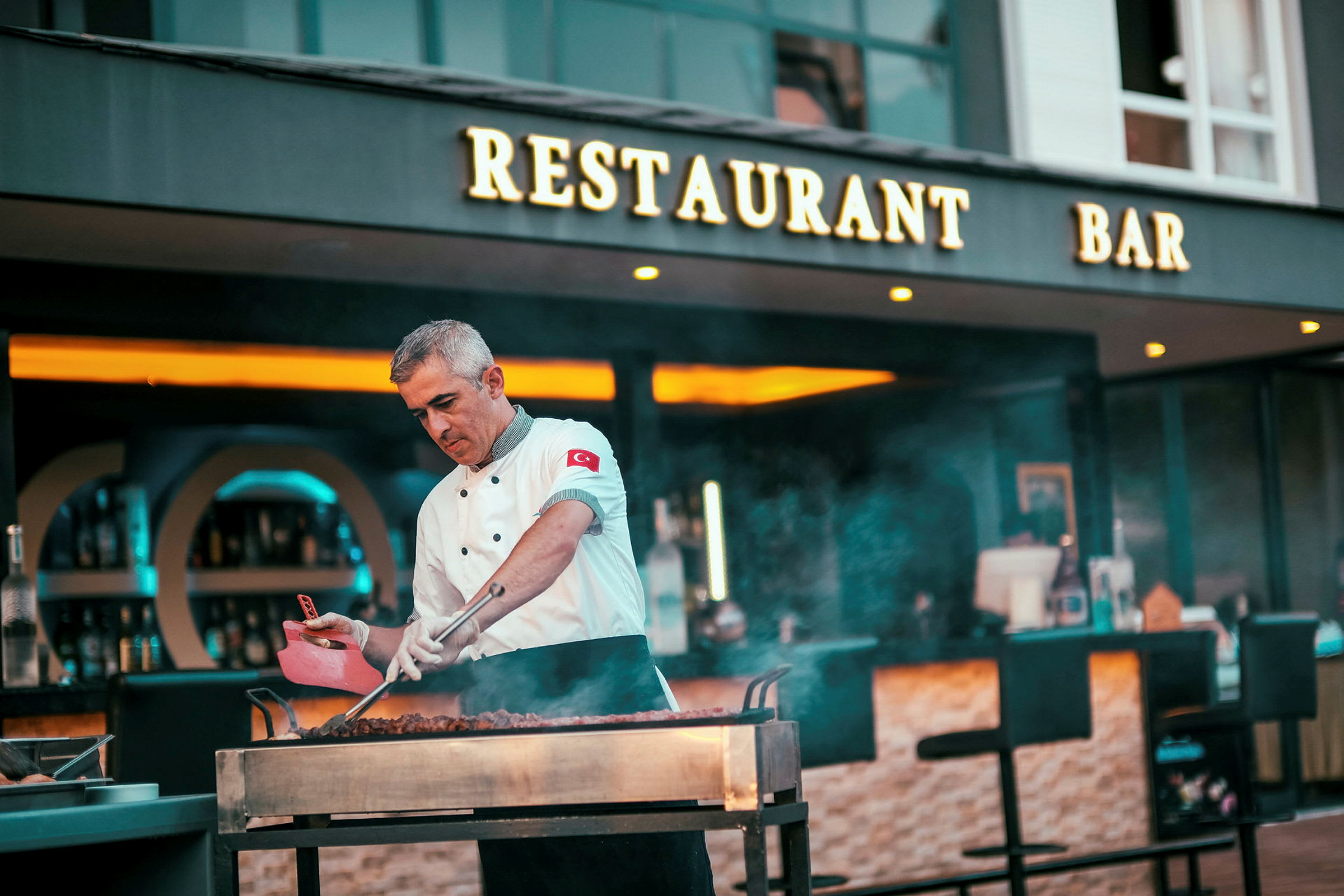 En kokk tilbereder mat på en åpen grill foran en restaurant og bar. (AI-generert tekst)