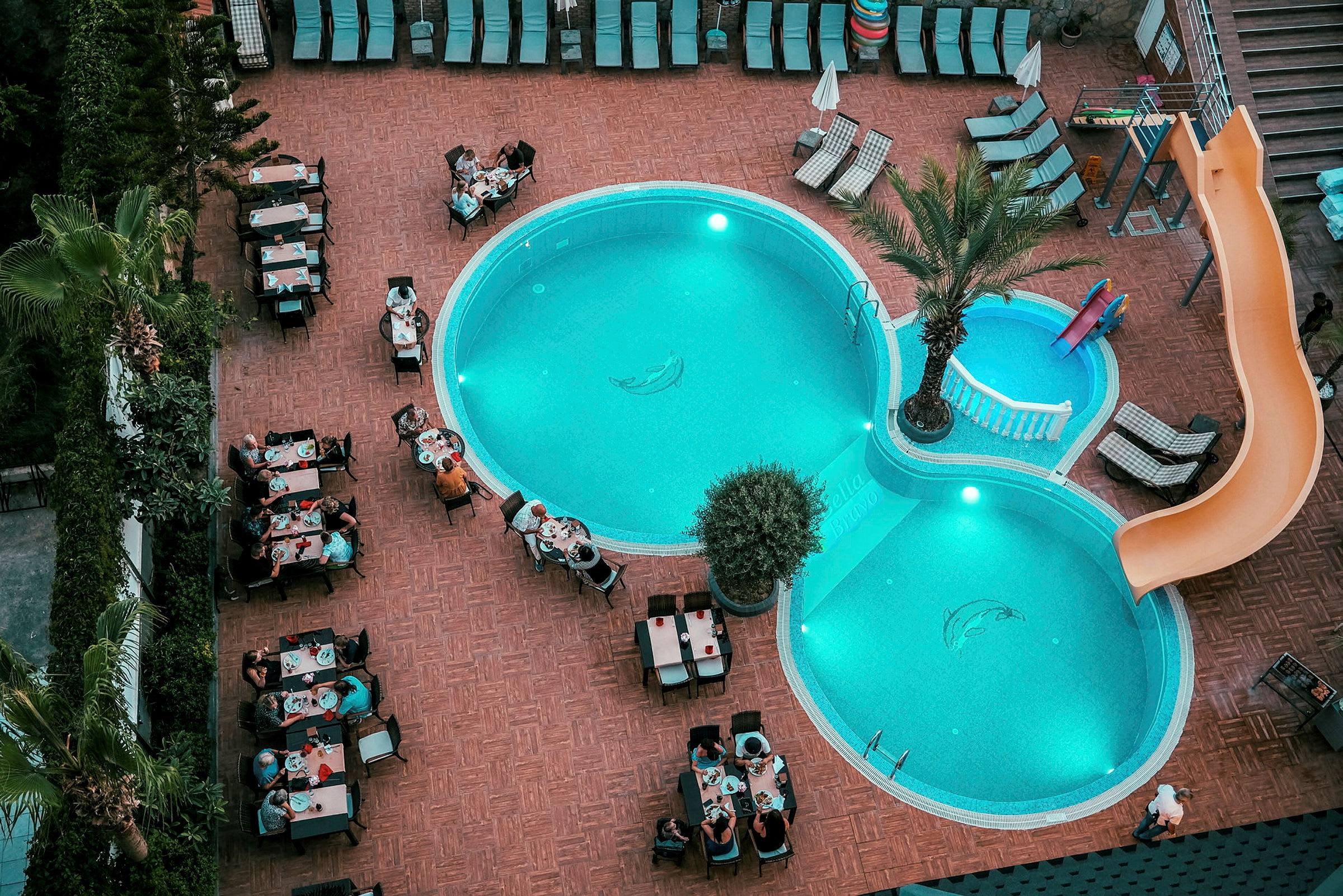 Et utendørsbasseng på et hotellområde med solsenger og personer som sitter ved bord og spiser. (AI-generert tekst)