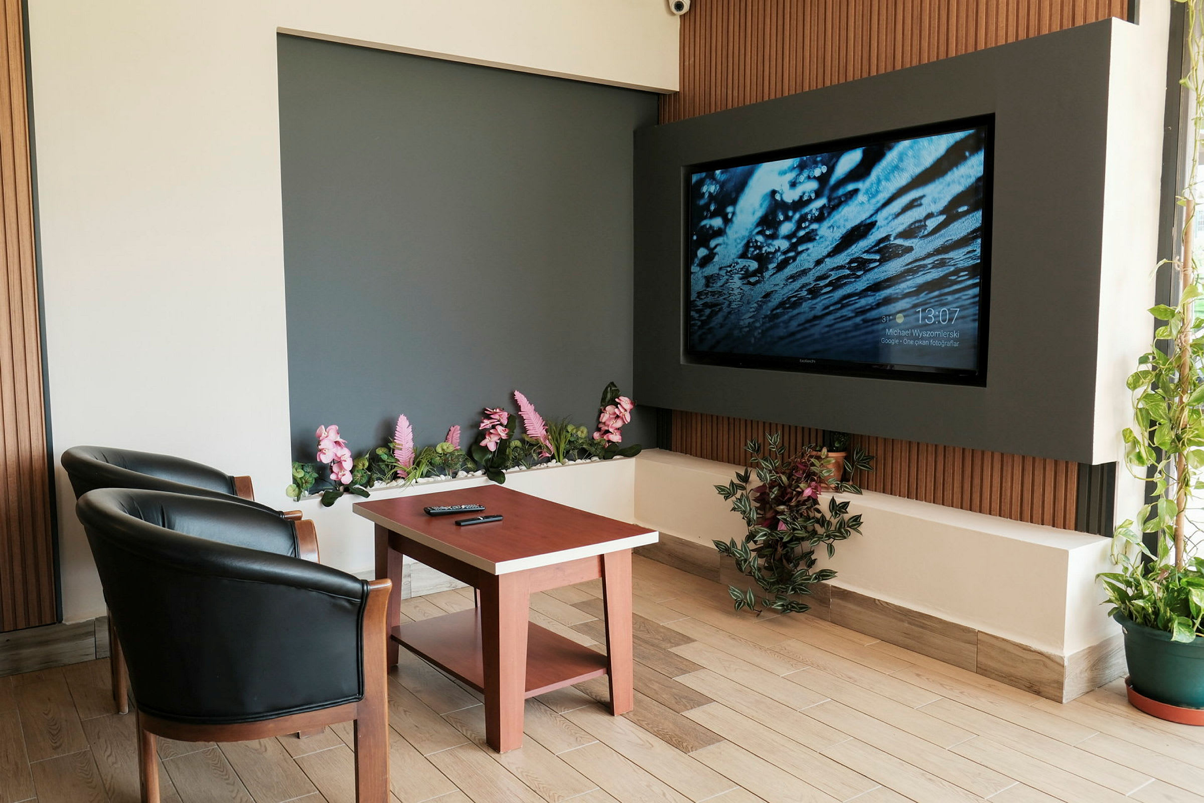 Et rom med to svarte lenestoler, et trebord og en stor veggmontert TV. Det er dekorative blomster og planter rundt. (AI-generert tekst)