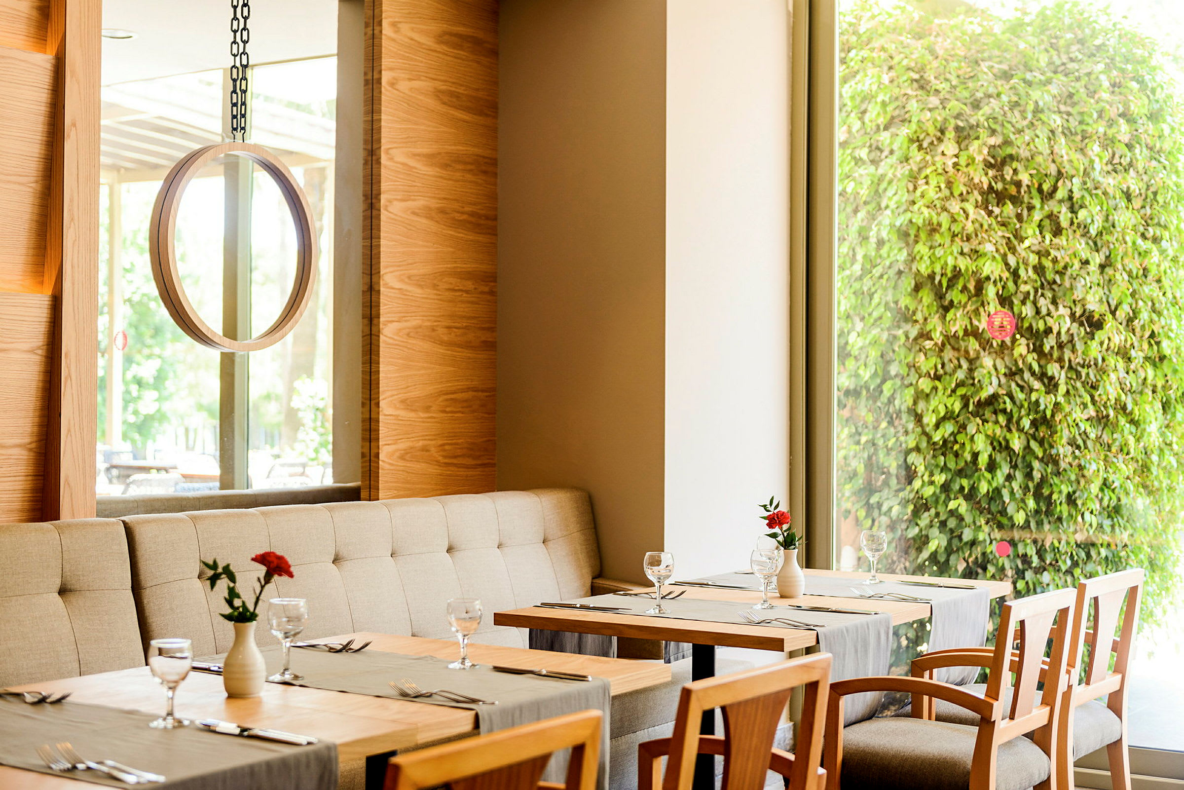 Et restaurantinteriør med bord, stoler og en grønn plantedekorasjon sett gjennom et vindu. (AI-generert tekst)