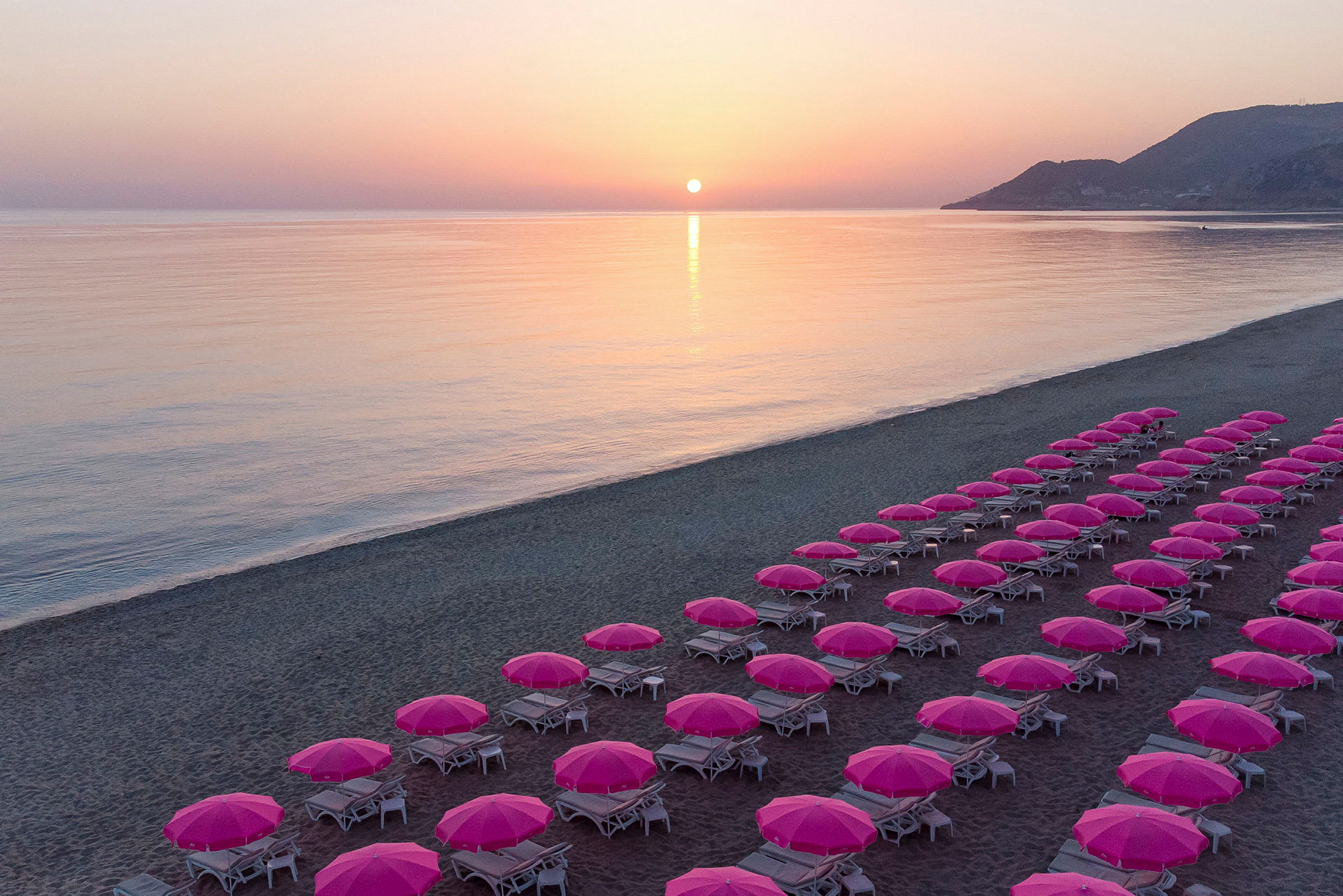 En strand ved solnedgang med rekker av solstoler under rosa parasoller. (AI-generert tekst)