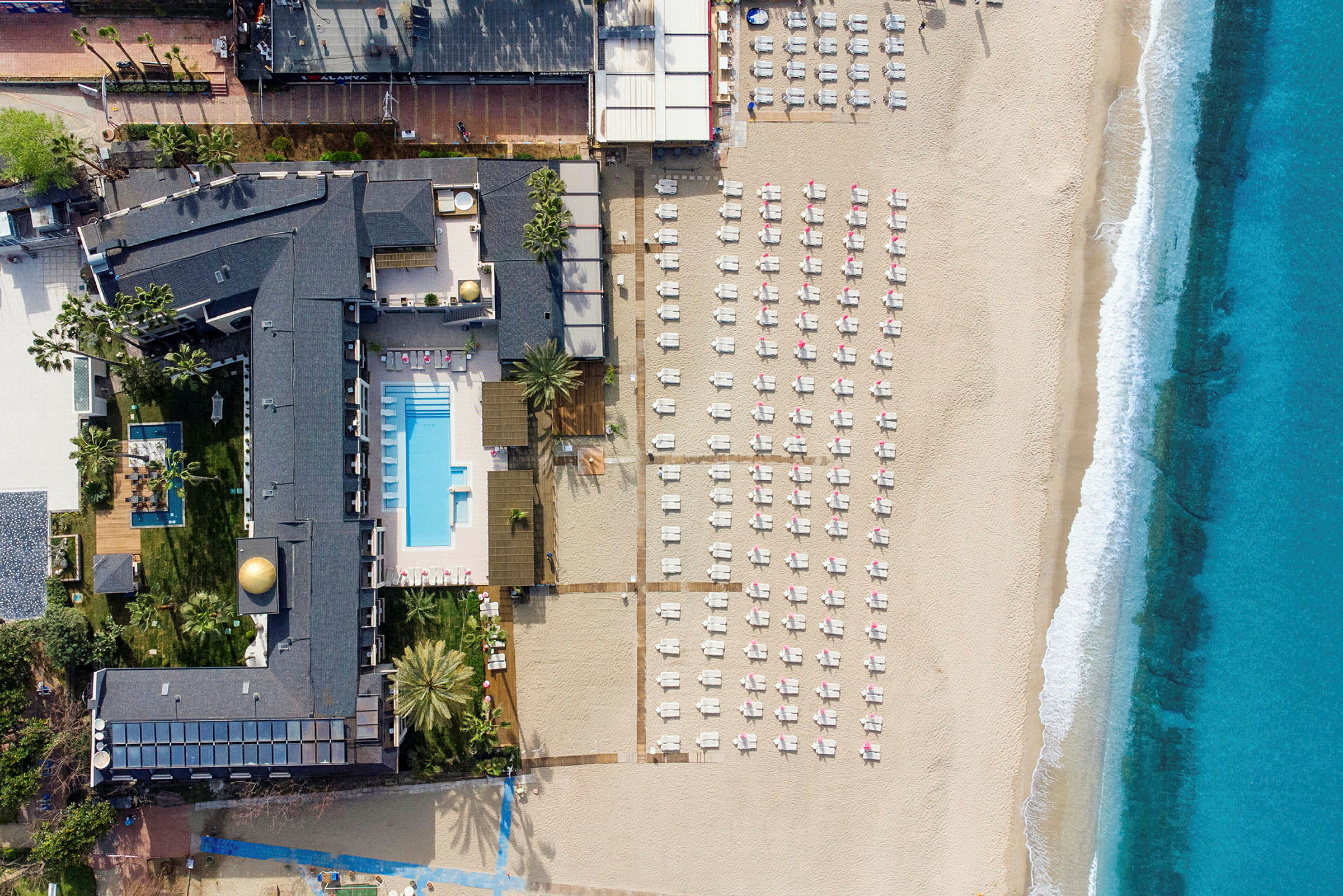 Luftbilde av en strand med solsenger, et hotellområde med basseng og havet. (AI-generert tekst)