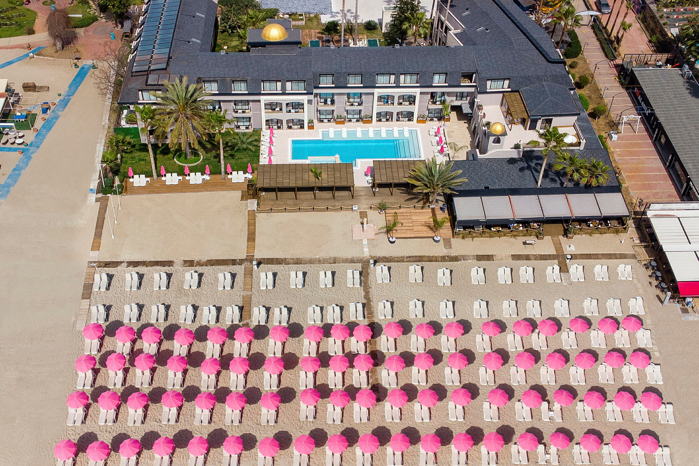 Fugleperspektiv på hotellområde med basseng og solsenger under rosa parasoller på stranden. (AI-generert tekst)
