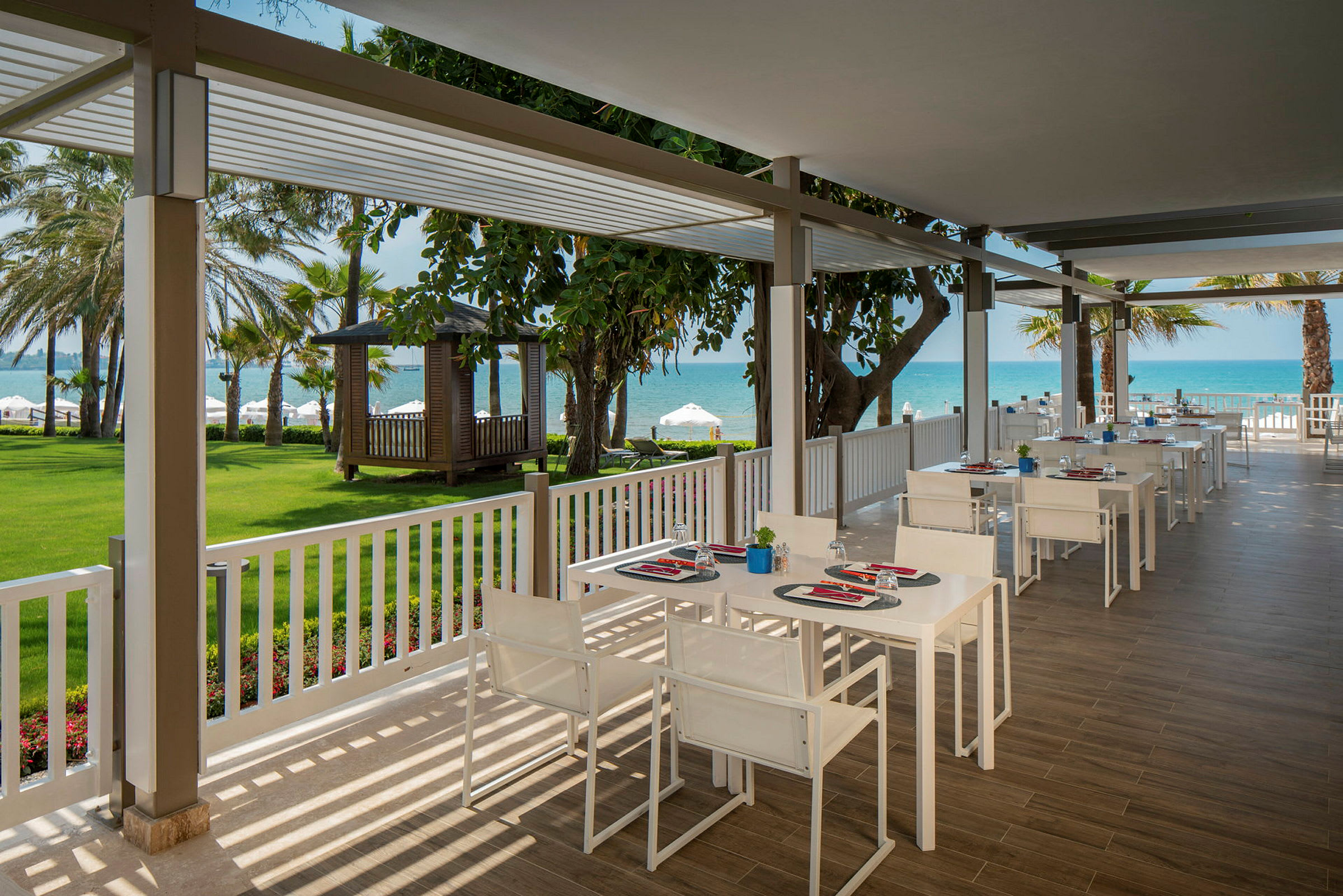 En utendørsrestaurant med hvite bord og stoler ved havet. Gressplen og trær i bakgrunnen. (AI-generert tekst)