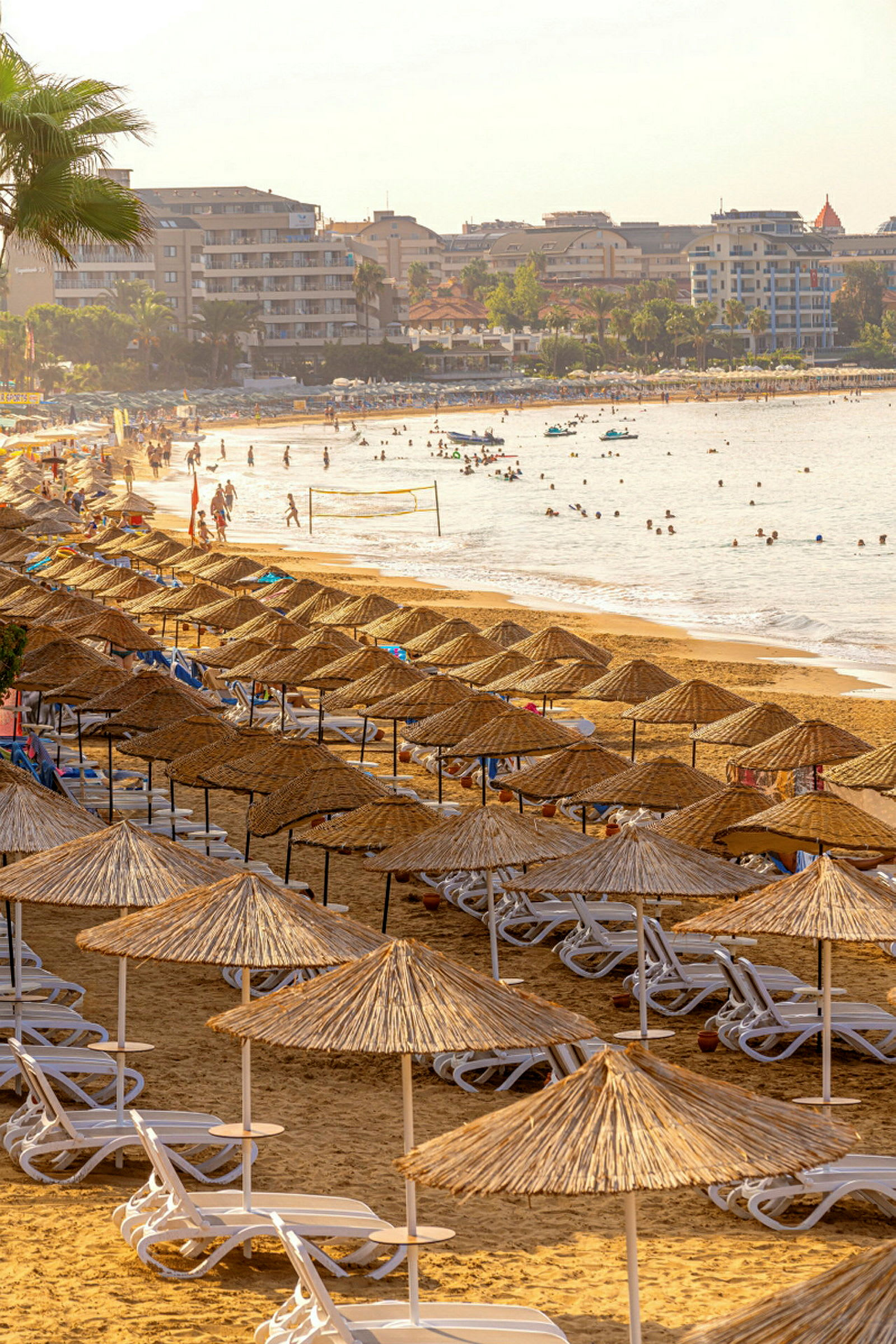 Strand med parasoller og solsenger i forgrunnen, bygninger og bading mennesker ved vannet i bakgrunnen. (AI-generert tekst)