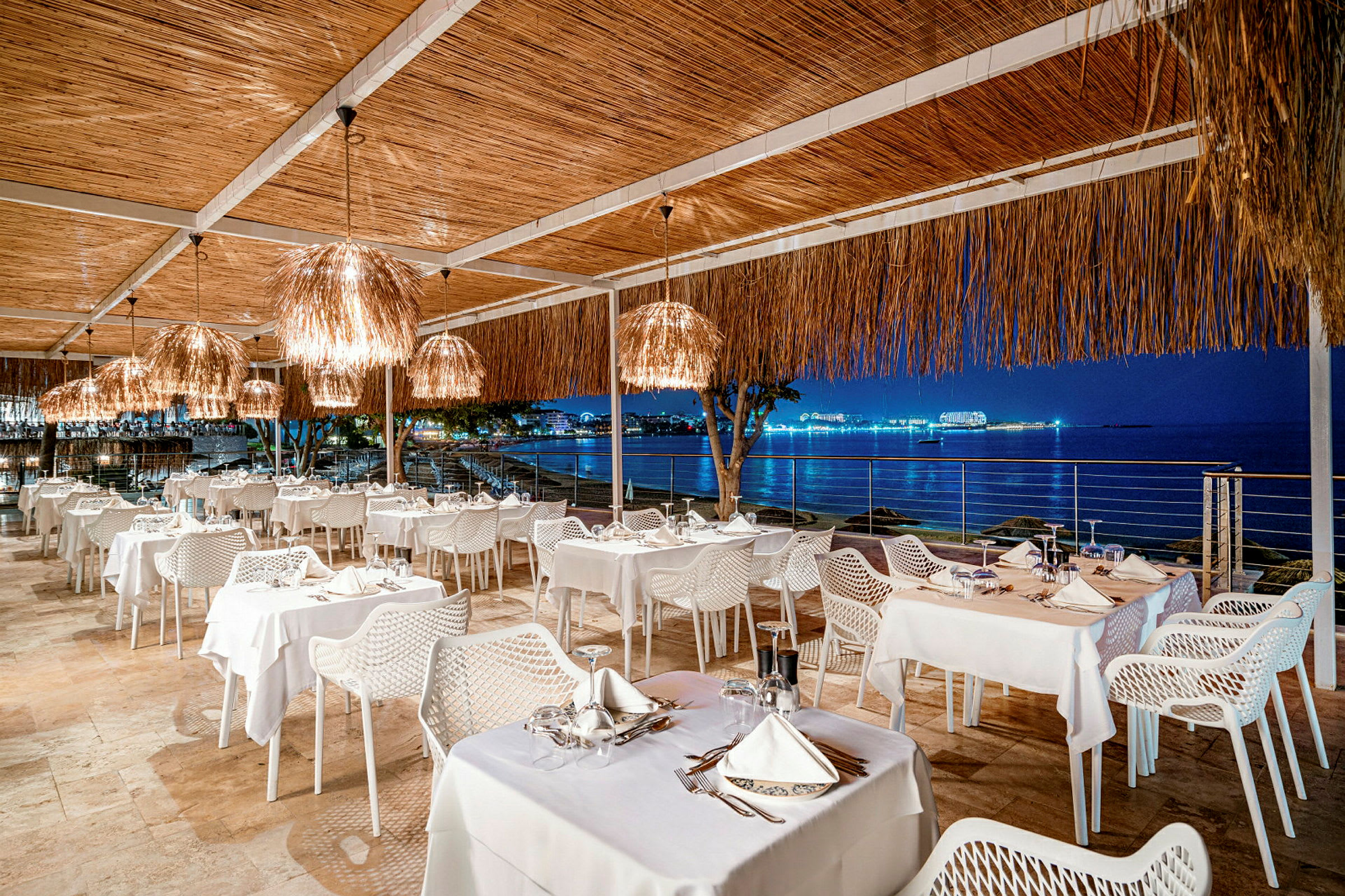 En utendørs restaurant med bord utstyrt med hvite duker og stoler, og utsikt mot en opplyst havbukt om natten. (AI-generert tekst)